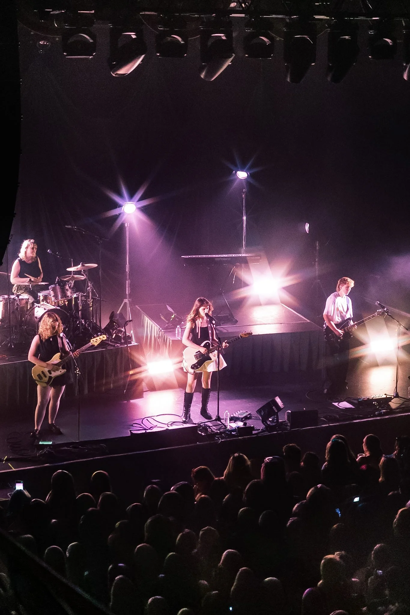 A live band performing on stage with musical instruments and purple stage lighting, audience visible in the foreground.