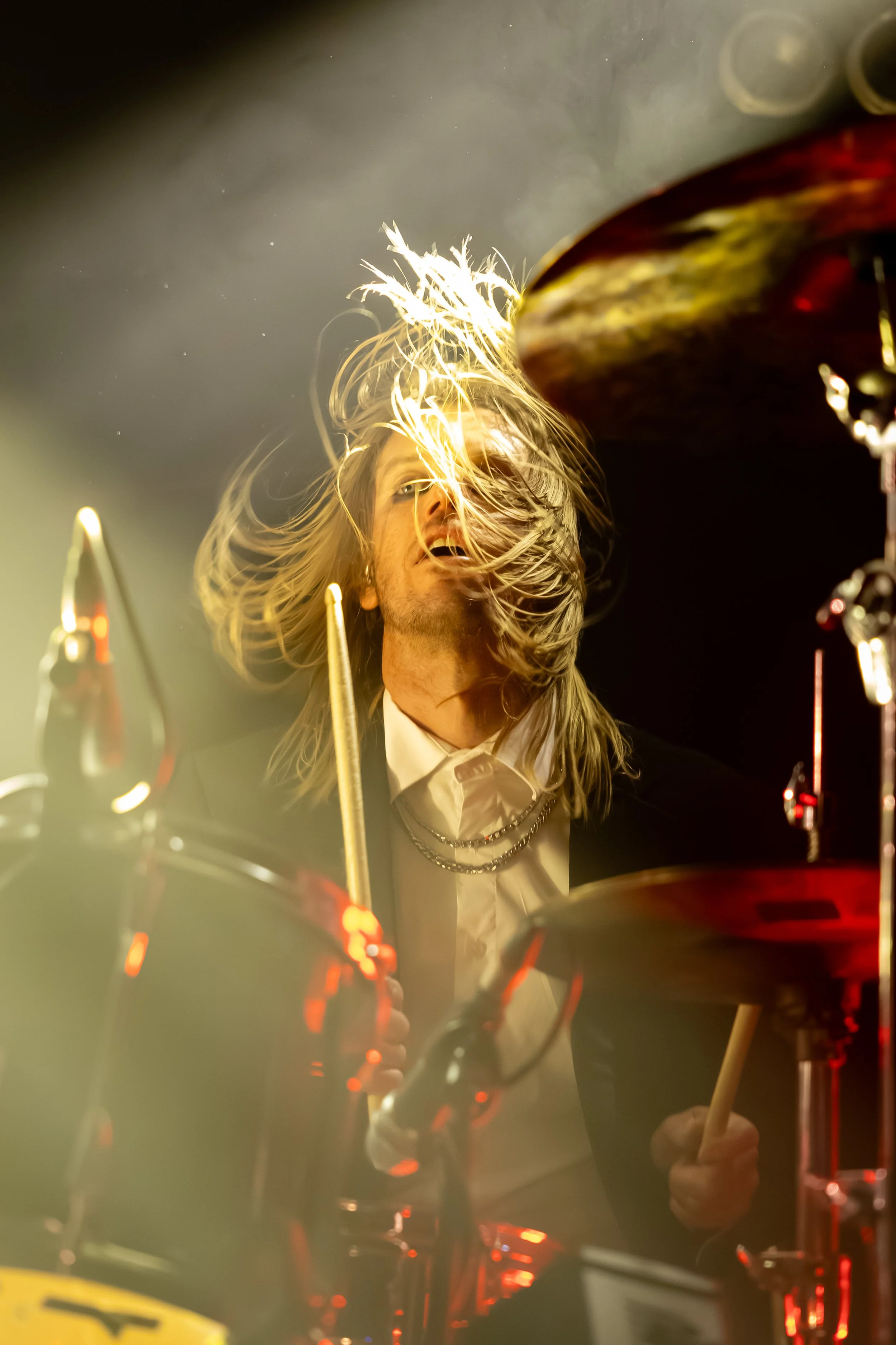 A drummer with long blonde hair playing drums, wearing a black blazer and white shirt, with drumsticks in hand, in a dimly lit setting with yellow and red lighting effects.