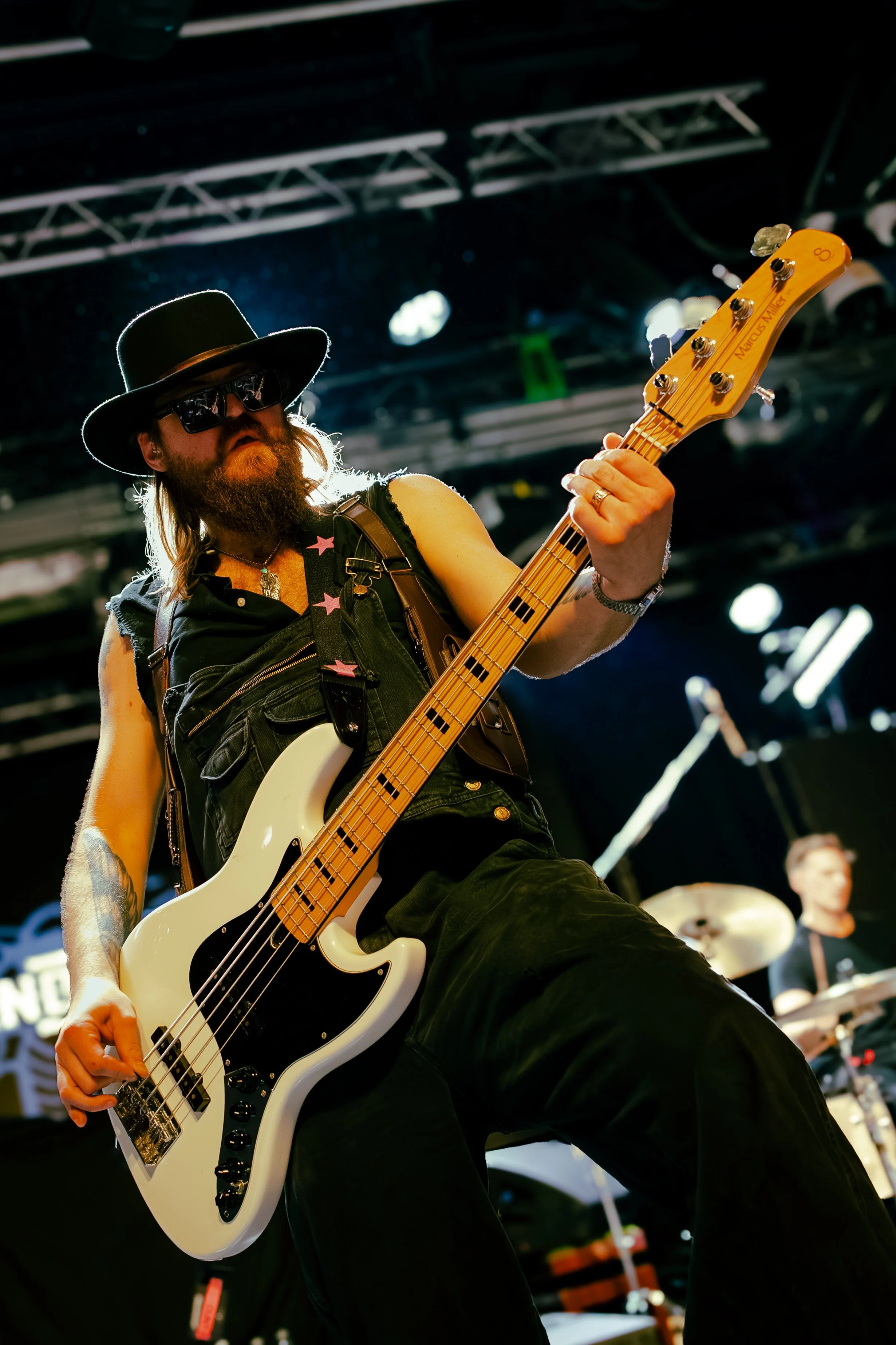 Musician with long hair, beard, sunglasses, and a wide-brimmed hat playing a white electric bass guitar on stage, with stage equipment and a drummer in the background.
