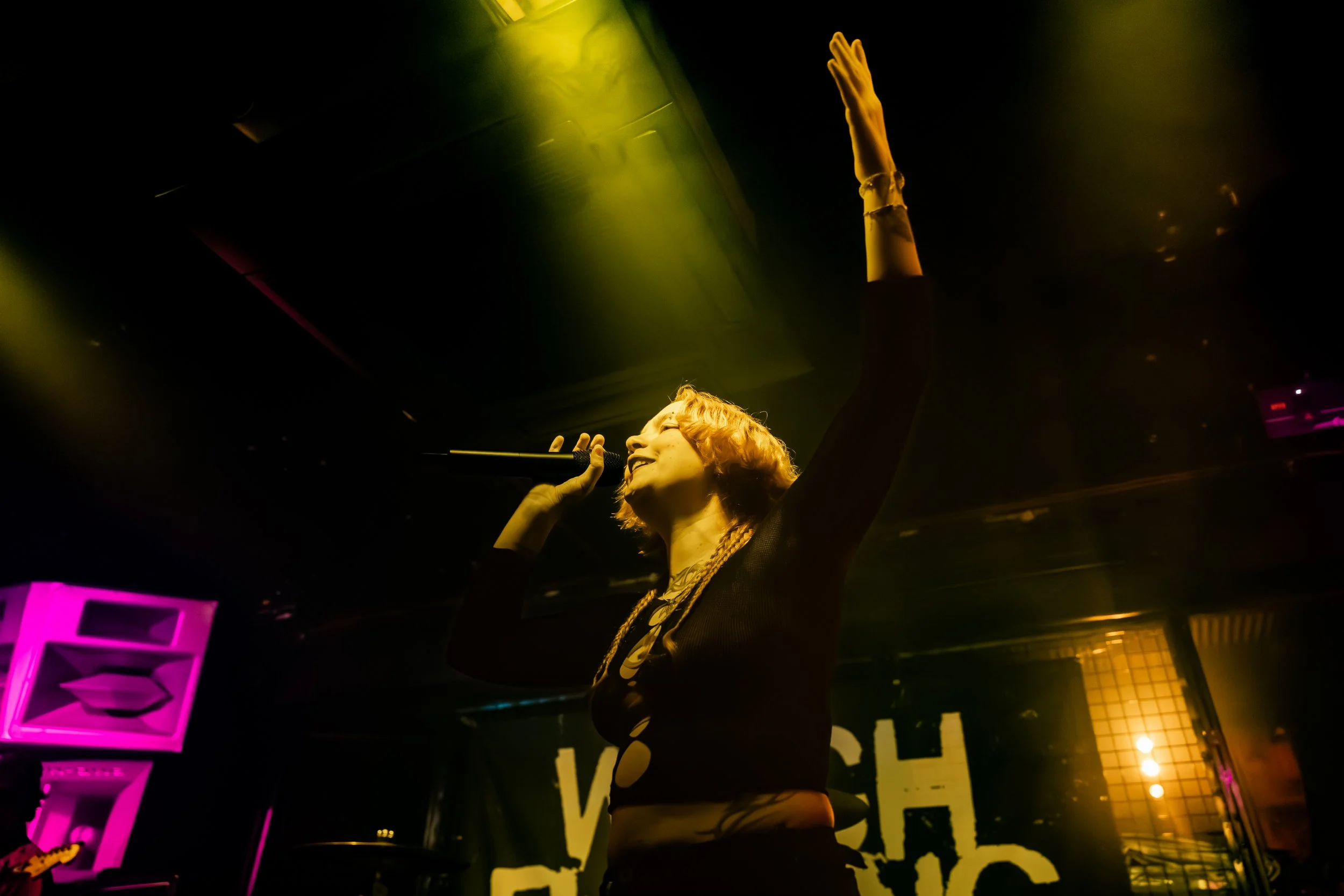 A woman singing or speaking into a microphone on stage with her right arm raised, surrounded by green and yellow lighting, in a nightclub or performance venue.