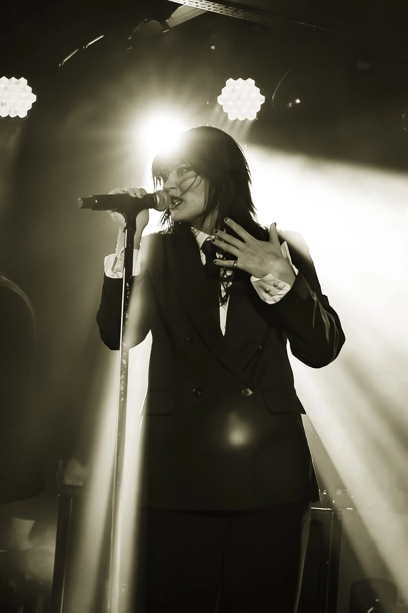 A woman with dark, wet hair singing into a microphone on stage, wearing glasses and a formal black suit with a white shirt, with bright stage lights creating a backlit effect.
