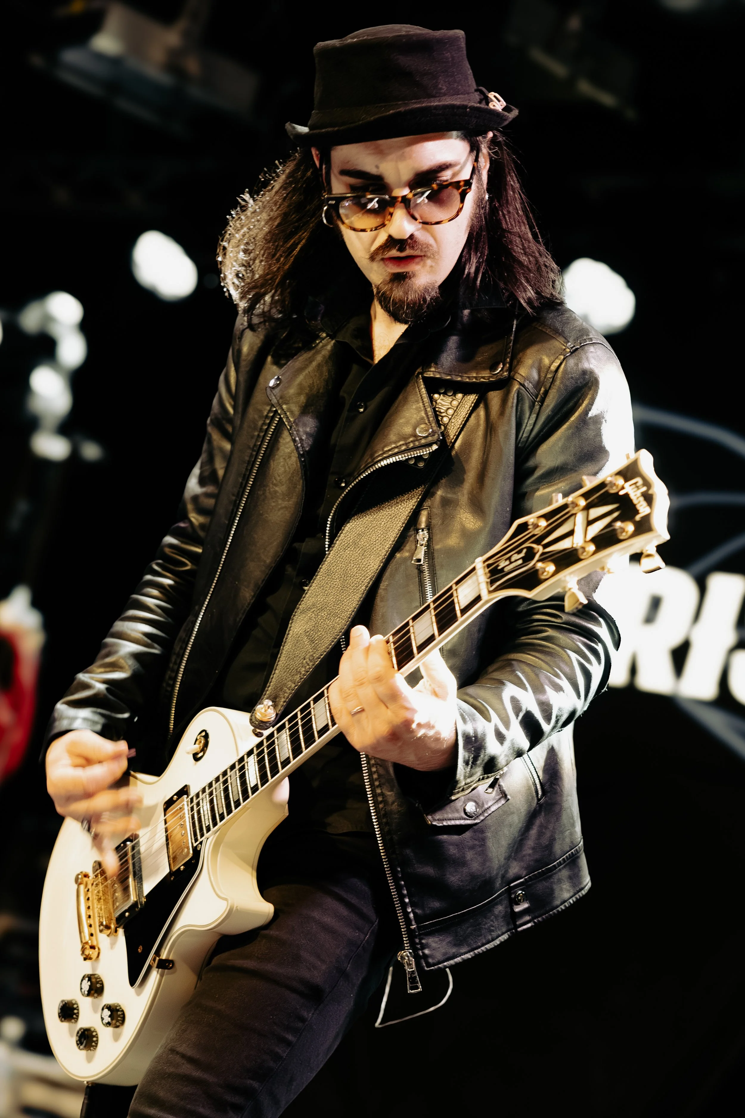A musician playing an electric guitar on stage, wearing a black leather jacket, black pants, sunglasses, and a black hat, with long hair and facial hair, under stage lighting.