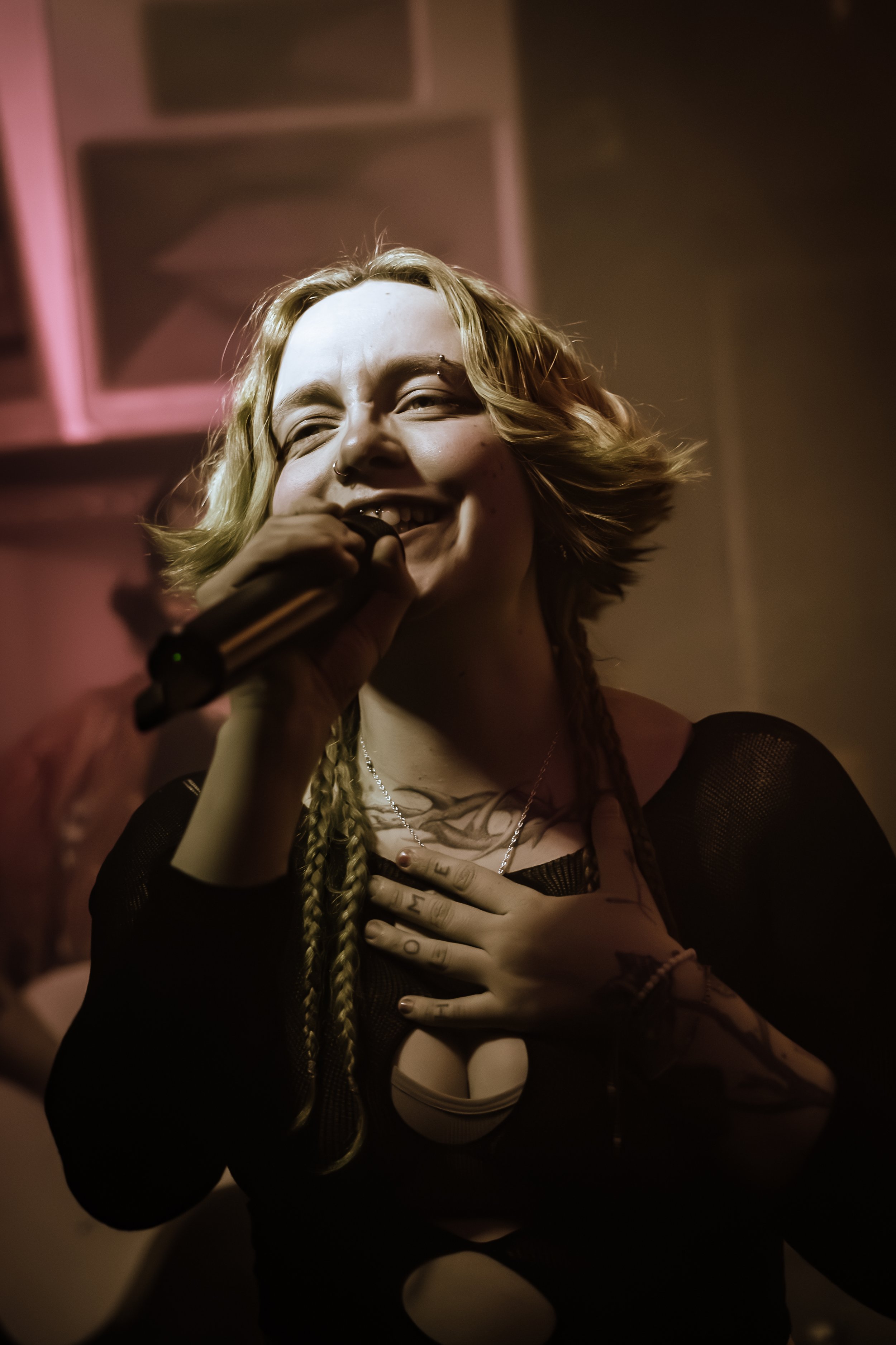 A woman with blonde hair, smiling, holding a microphone close to her mouth, with her hand on her chest, in a dimly lit room.