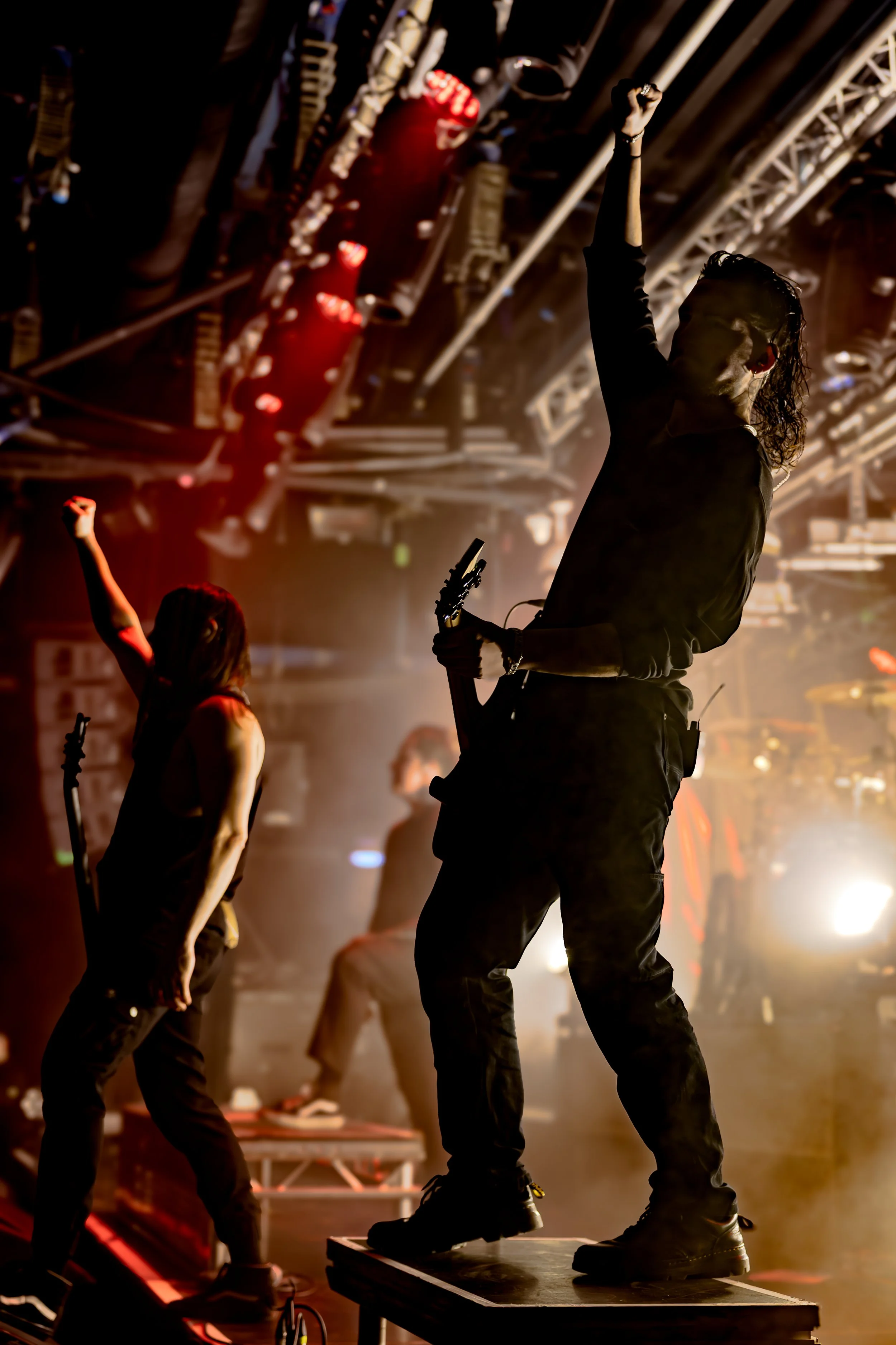 Musicians performing on stage with one person playing an electric guitar and others dancing or playing instruments, in a concert setting with stage lights and equipment.
