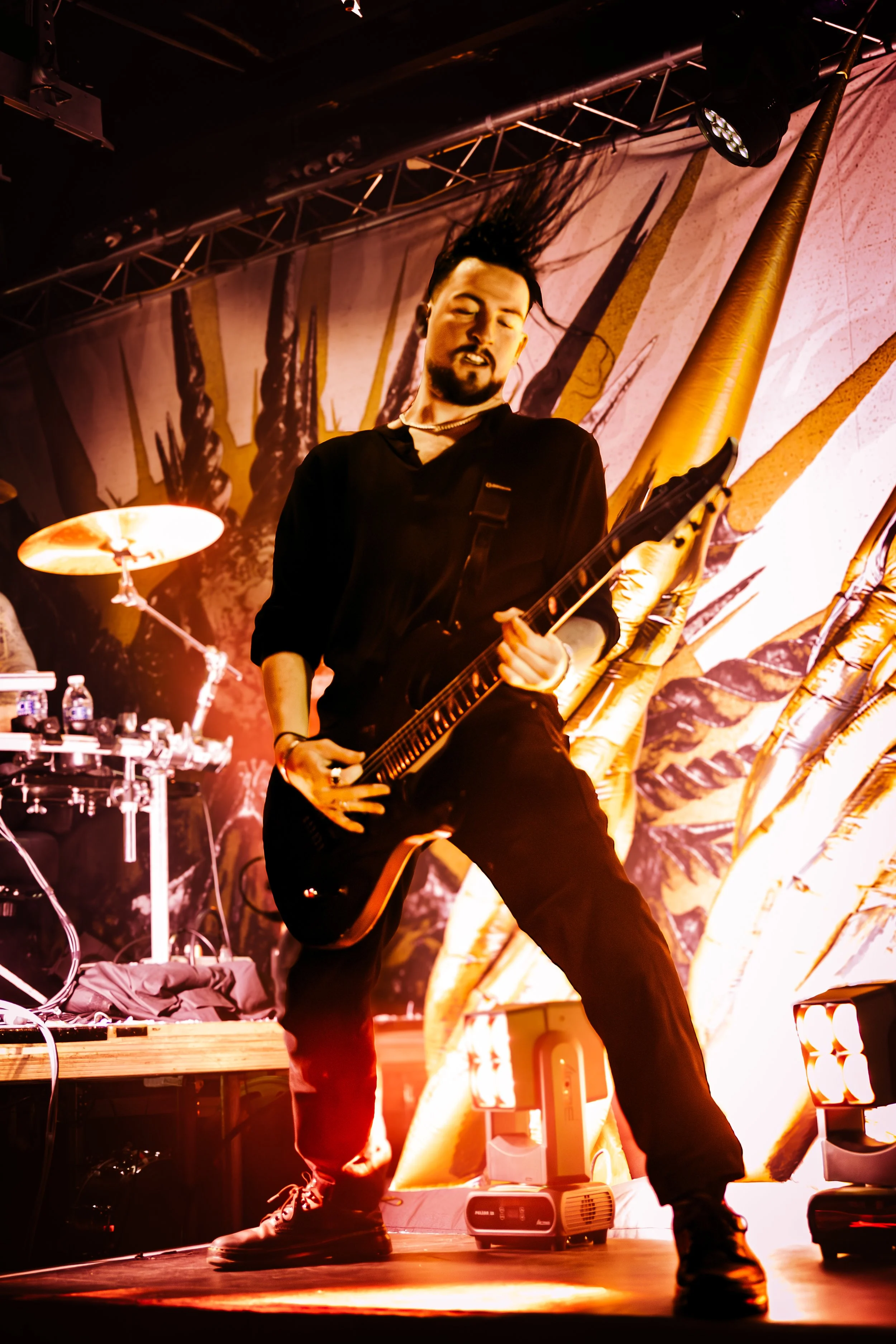 A male guitarist passionately performing on stage with long hair flying in the air, illuminated by warm stage lighting, and a backdrop featuring abstract artwork.