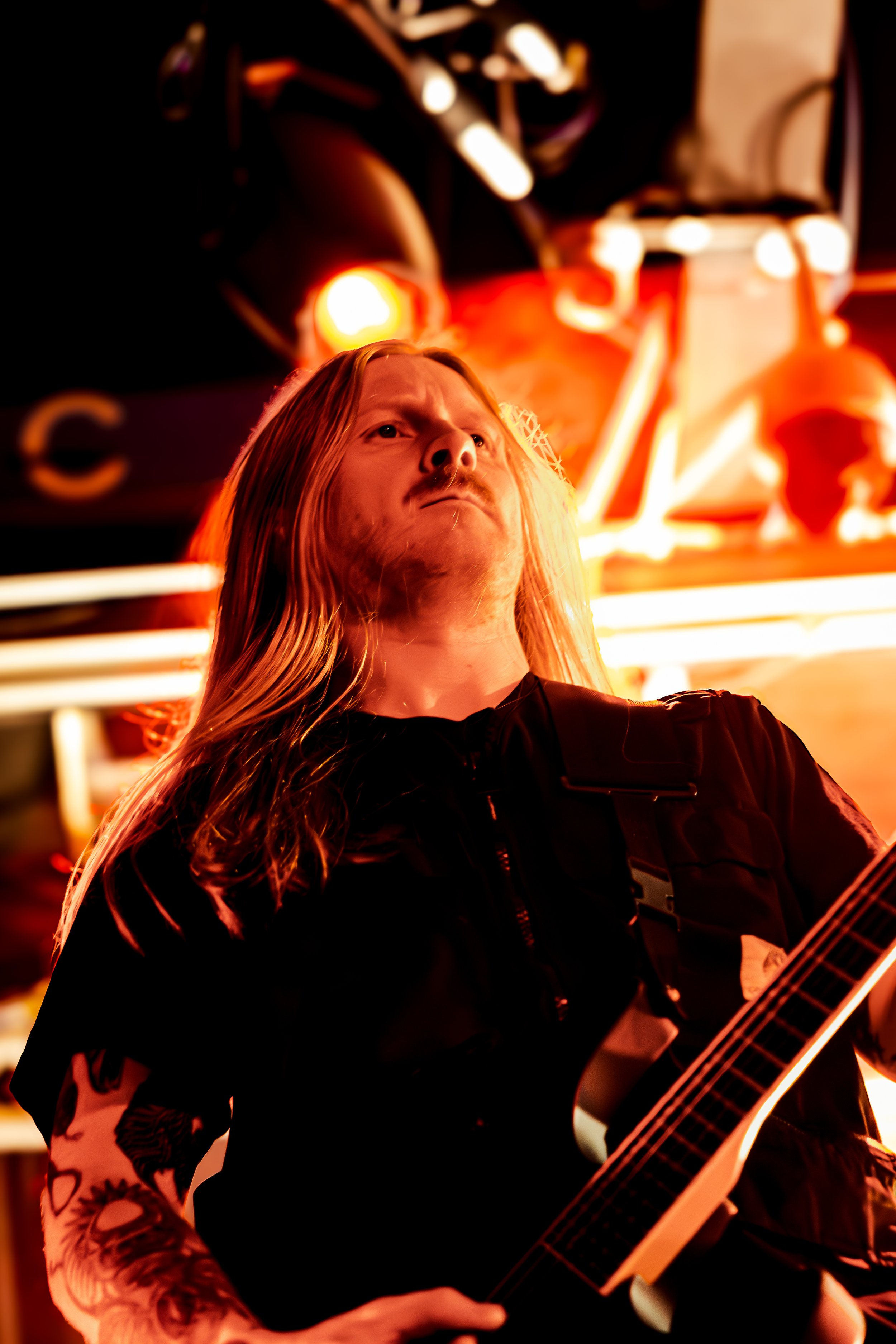 A man with long blond hair and tattoos on his arm, playing an acoustic guitar in a dimly lit setting with orange and yellow lights.