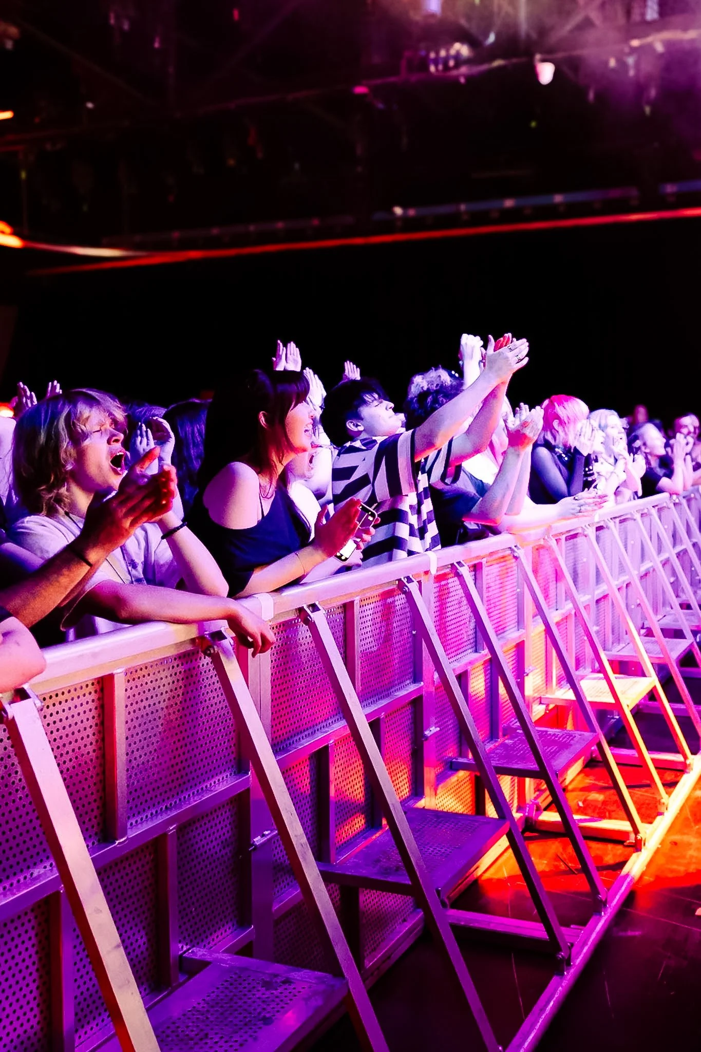 Concert crowd behind a metal barrier with pink and purple lighting.