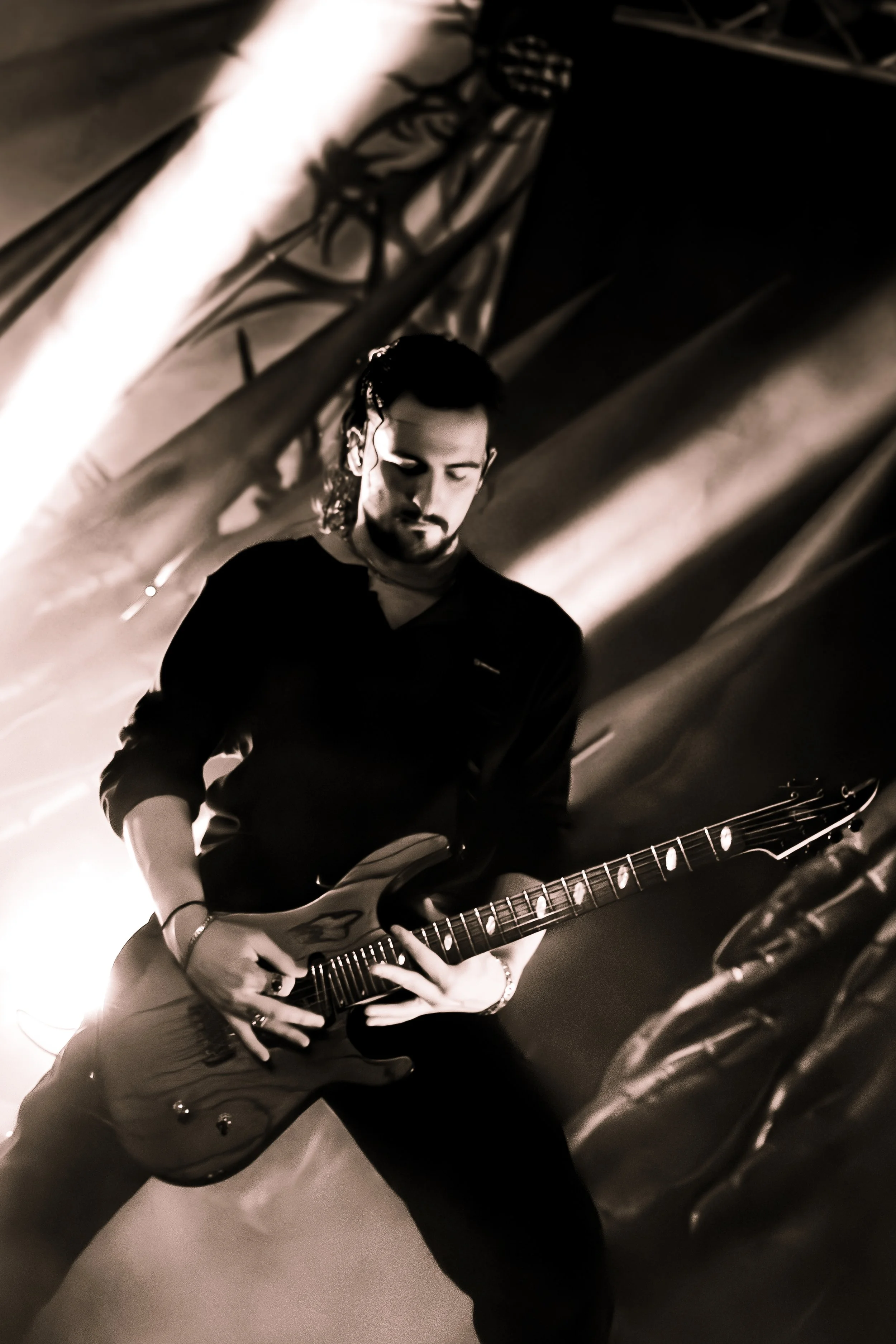 A man playing an electric guitar in a dimly lit room, with his eyes closed and wearing a dark shirt.