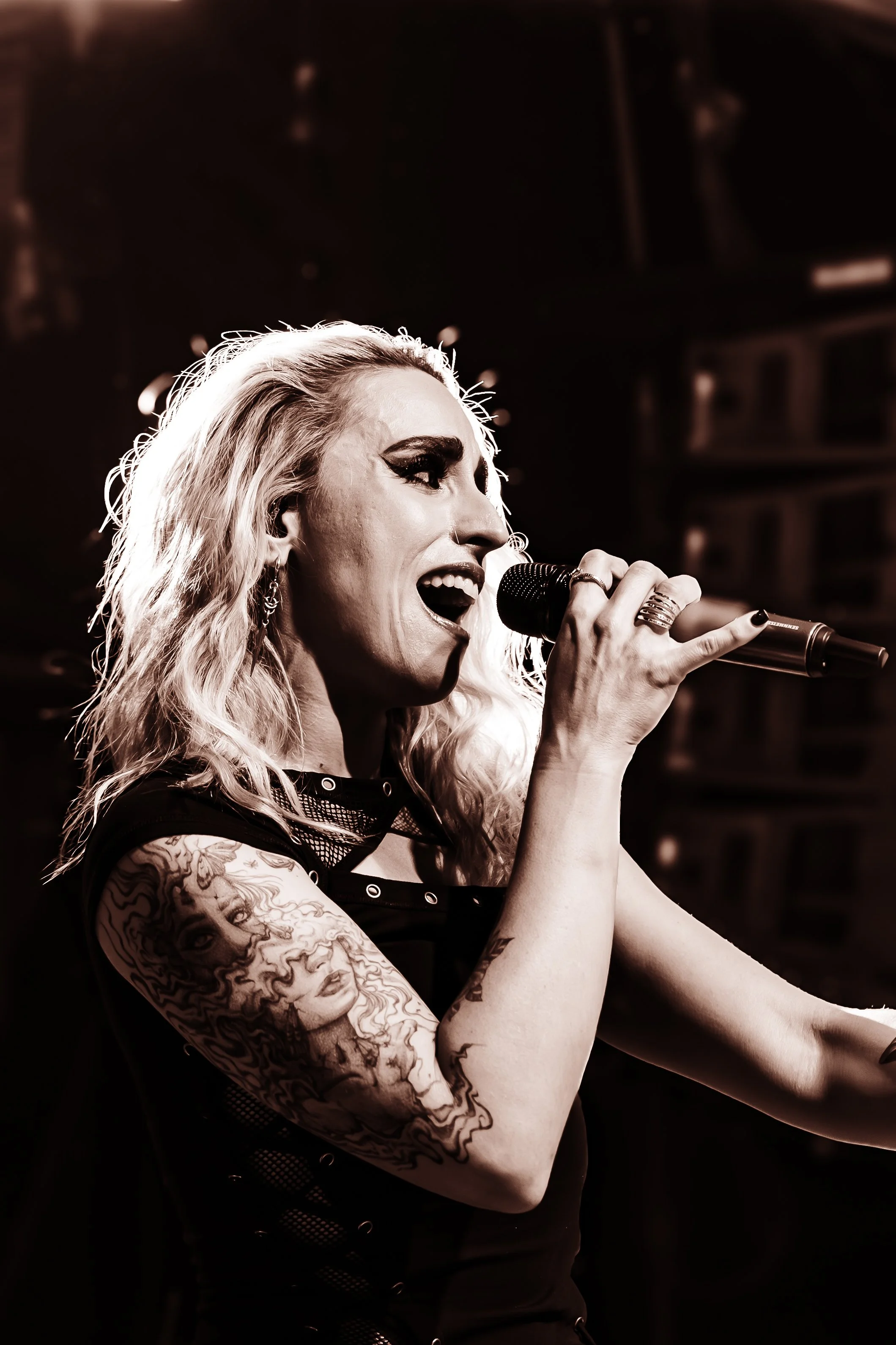 A woman with blonde wavy hair singing into a microphone, with tattoos on her arm, wearing makeup with dark eyeliner, in a dark venue.