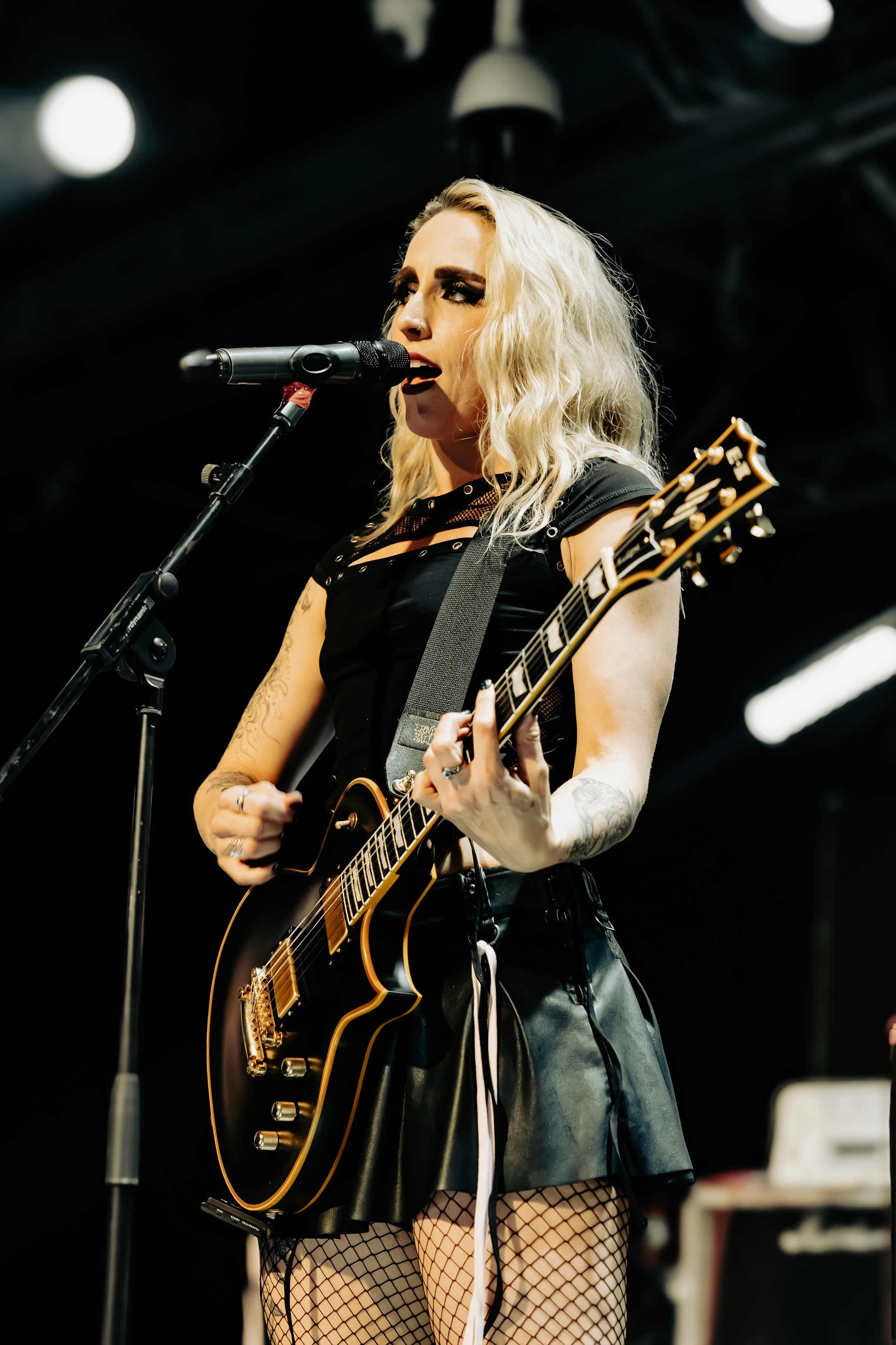 A woman singing into a microphone while playing an electric guitar, with platinum blonde hair, tattoos, a black outfit, fishnet stockings, and a dark stage background.