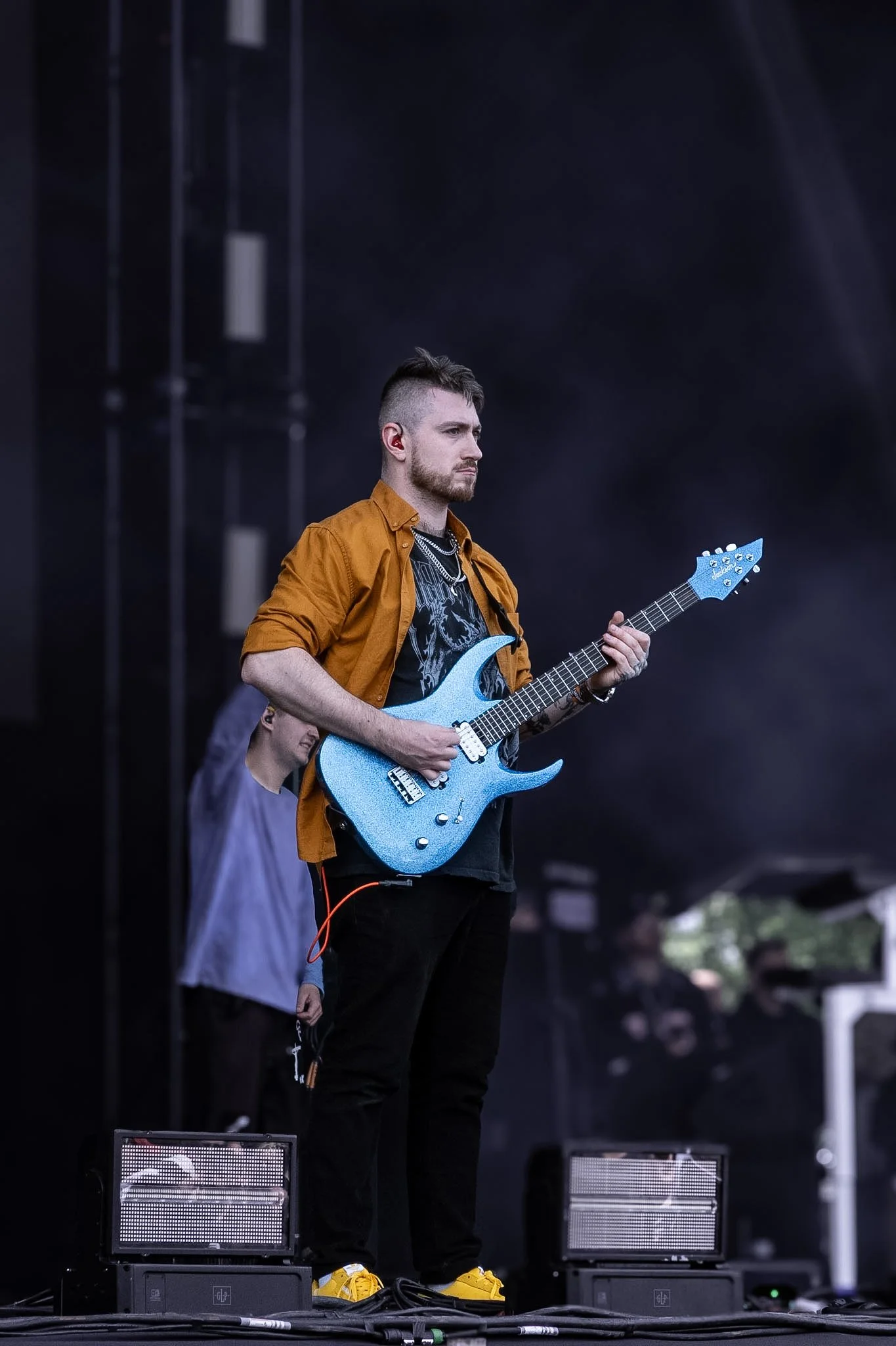 A musician with short hair and a beard playing a light blue electric guitar on stage, wearing an orange shirt, black pants, and yellow sneakers, with stage equipment and a dark background.