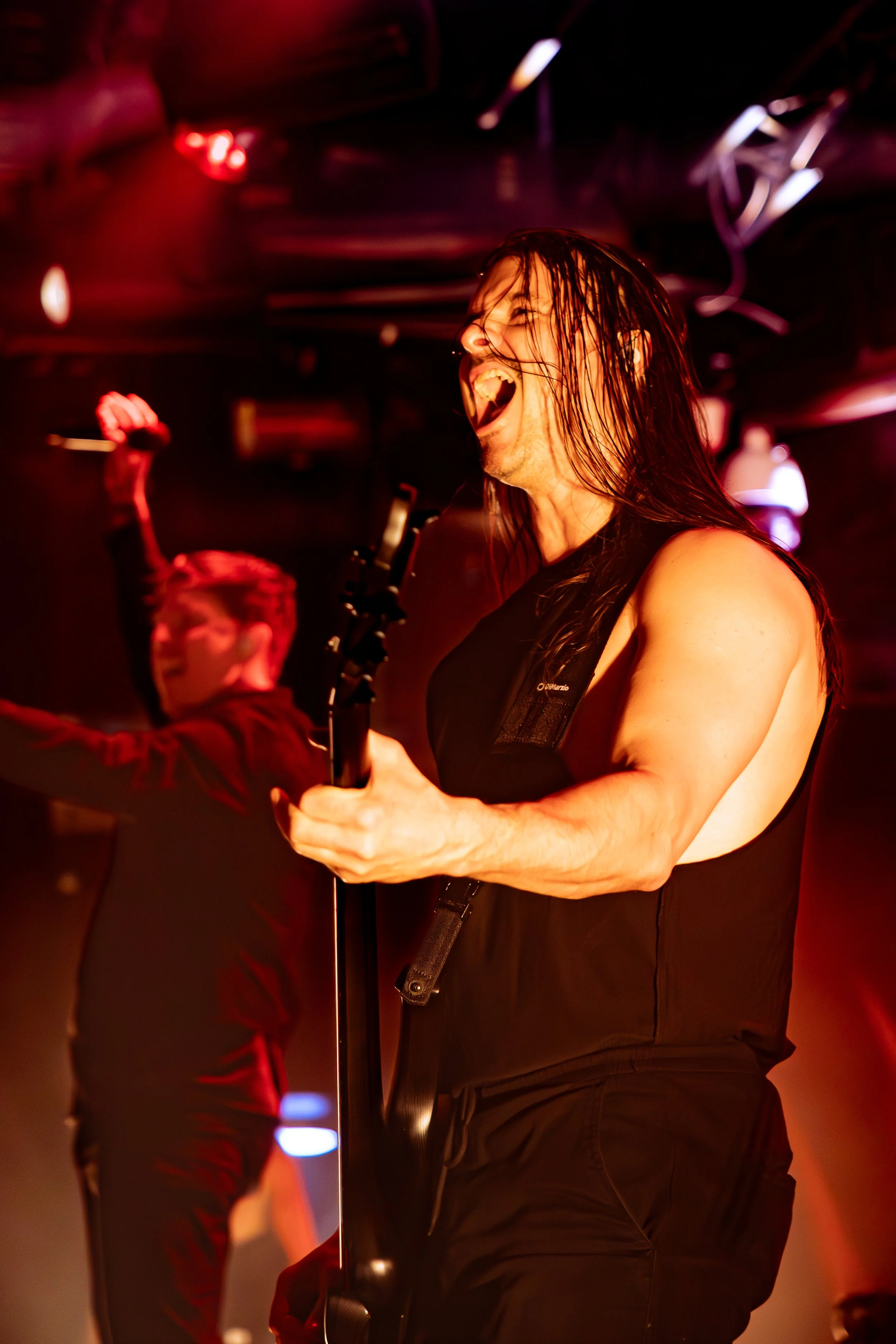 A musician singing passionately into a microphone on stage, with long wet hair, wearing a black outfit, red stage lighting.