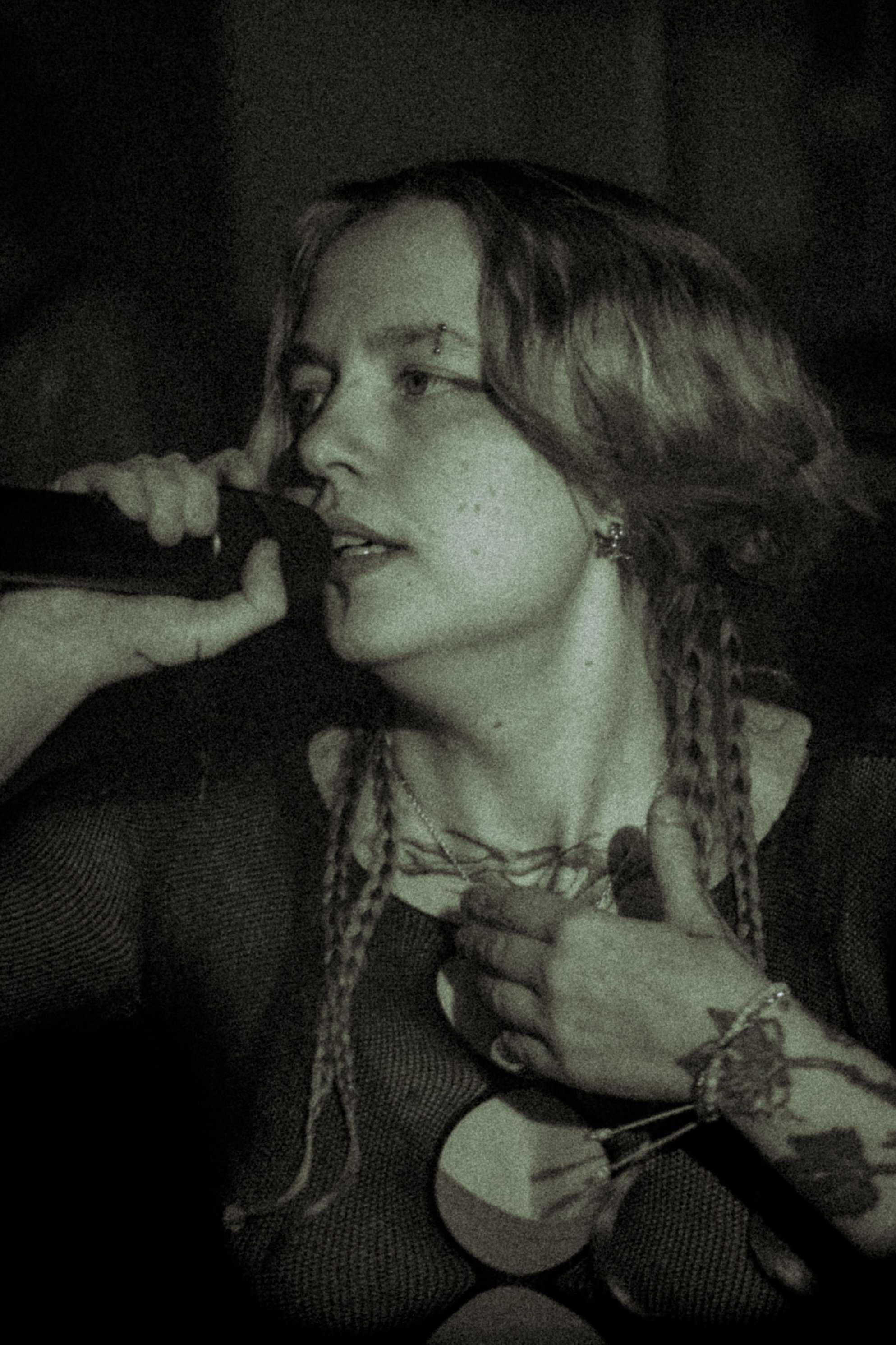 A person with long, wavy hair and nose piercing appears to be singing or speaking into a microphone in a dark setting. They are making a peace sign with one hand and holding a guitar with the other.