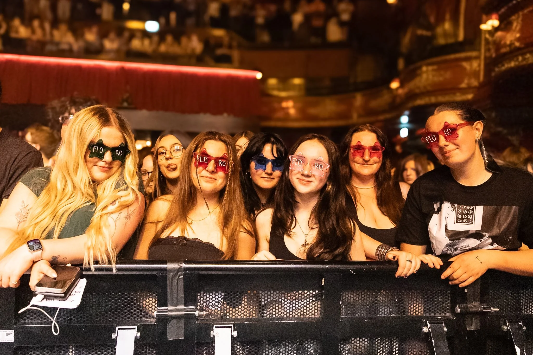 Group of young women wearing colorful 'FLO' and 'RO' glasses at a concert or event, standing behind a barrier with a dark, ornate theater interior in the background.