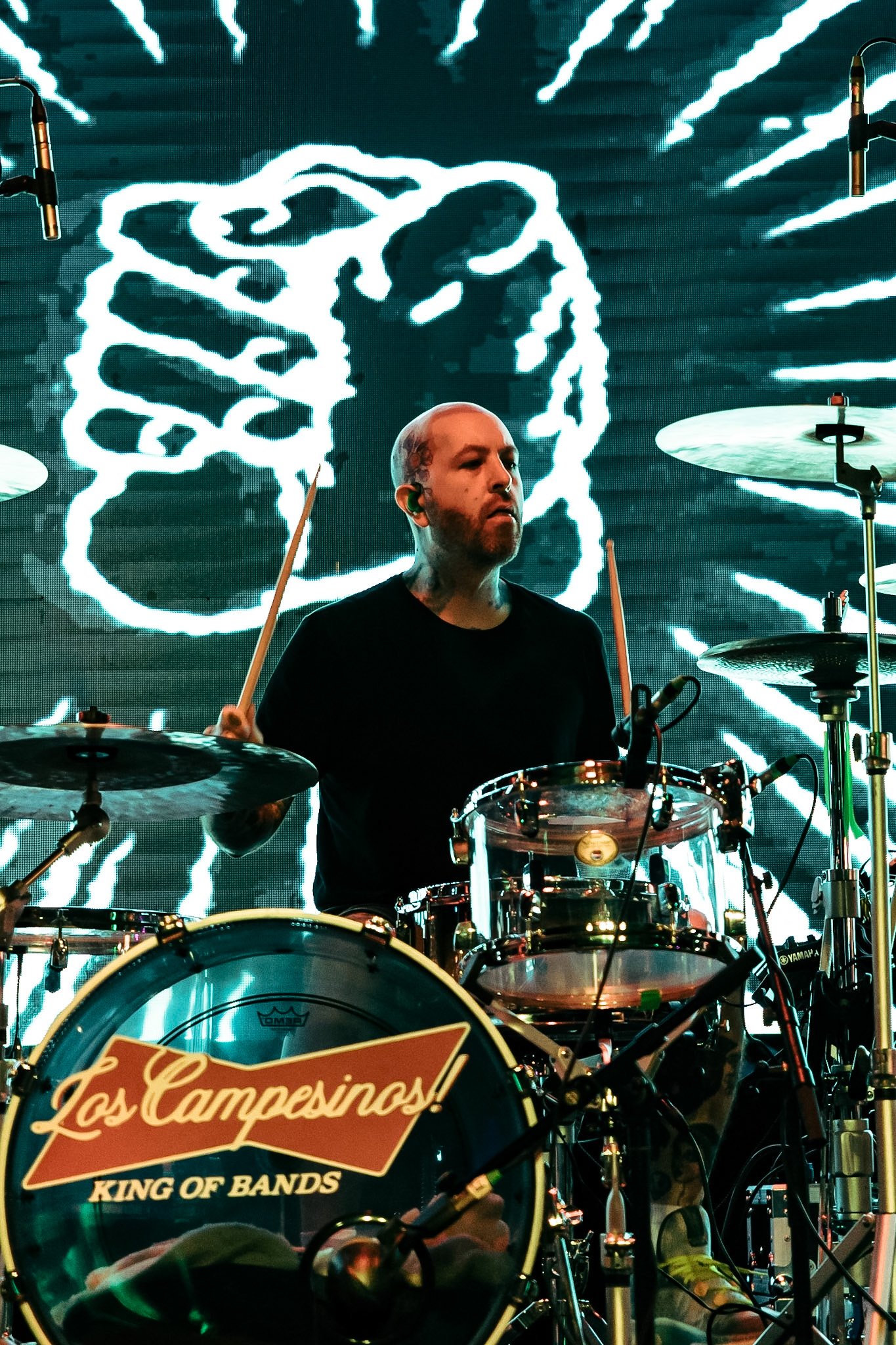 A drummer playing on stage with a large digital screen behind him displaying a stylized fist and lightning bolts. The drum set bears a logo reading "Los Camperos, King of Bands".