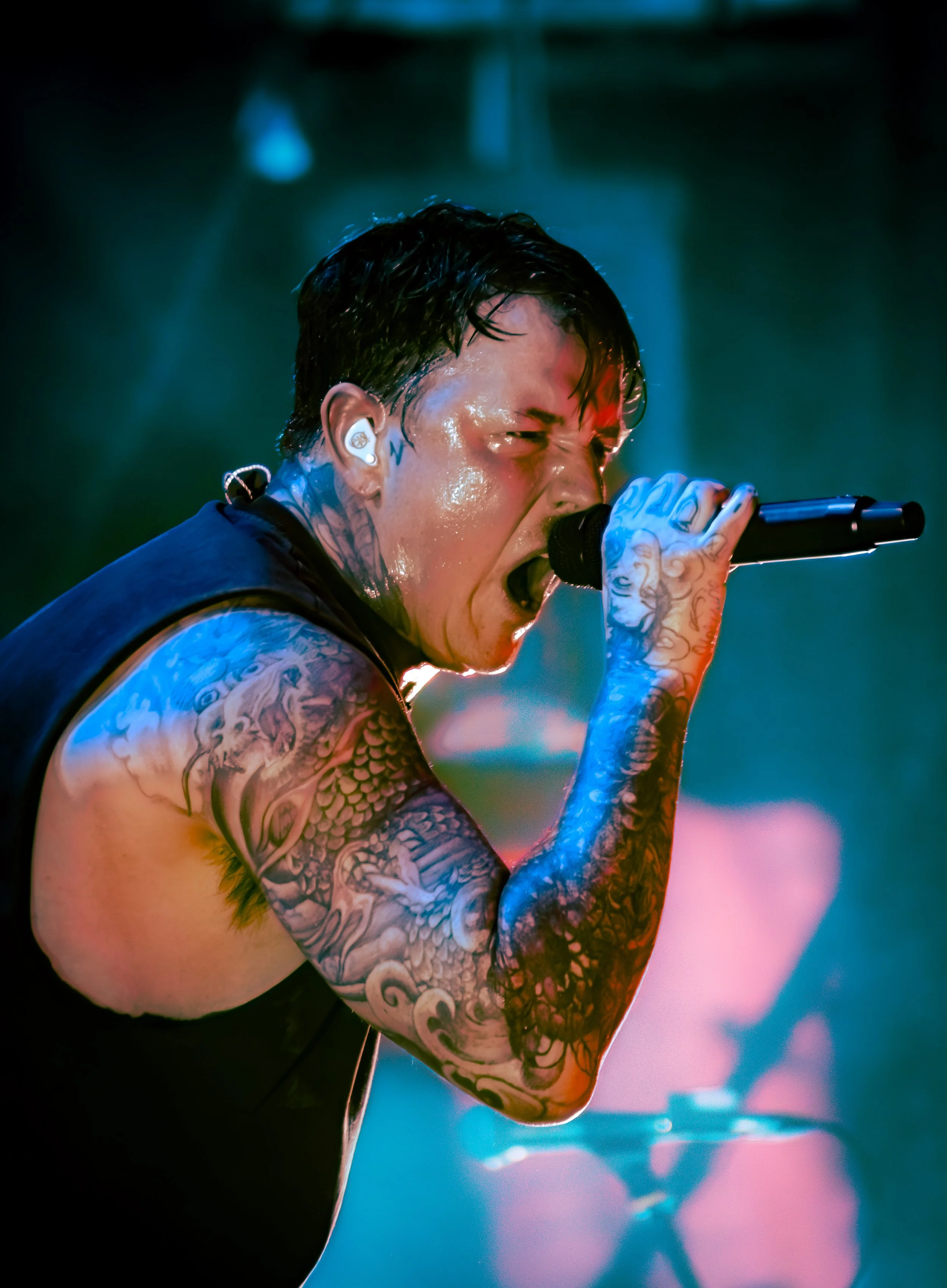 Close-up of a person with short dark hair singing into a microphone on stage, with tattoos on their arm and a leather sleeveless shirt