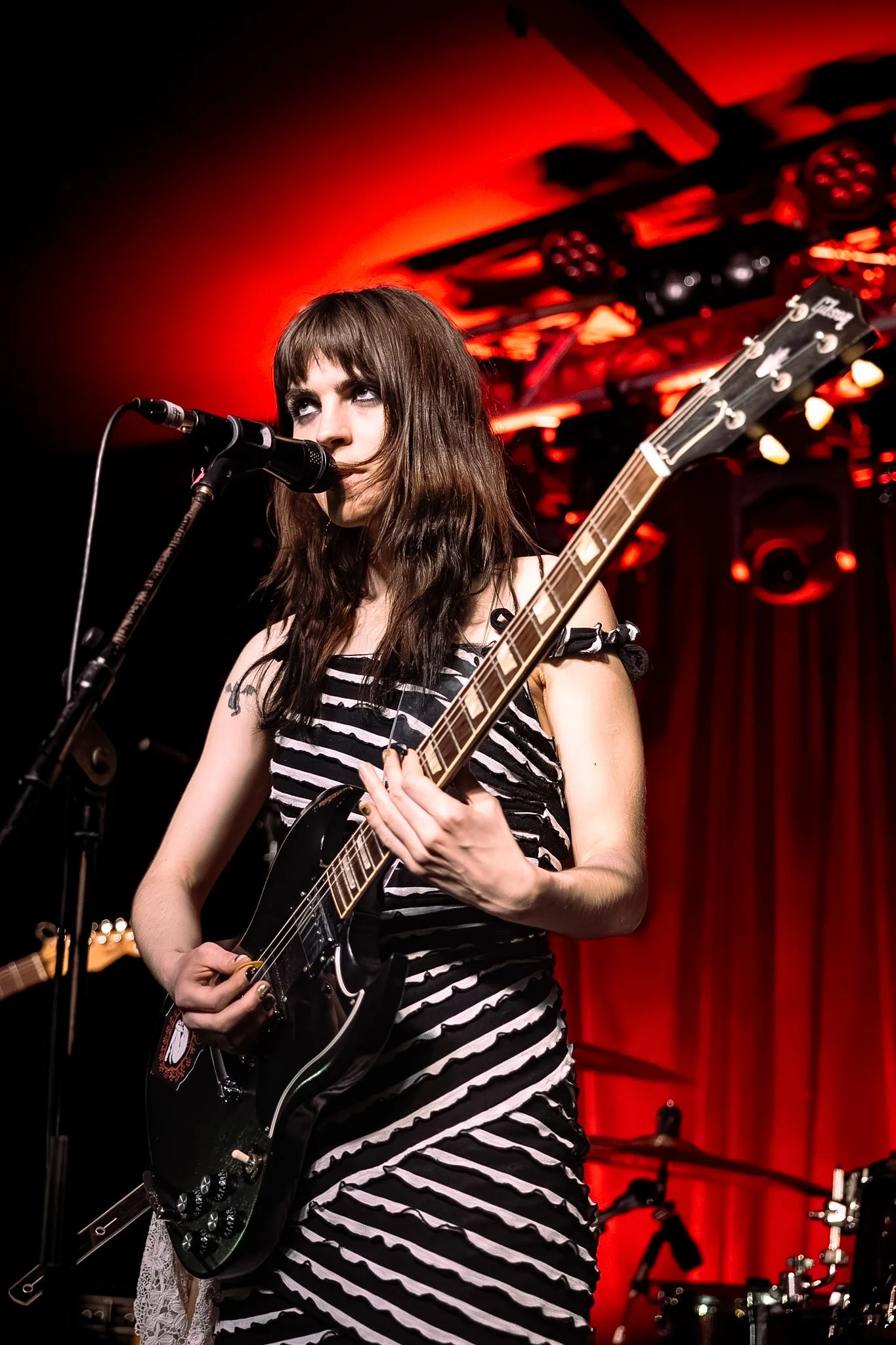 A young woman with long brown hair playing an electric guitar and singing into a microphone on stage with red lighting and concert equipment in the background.
