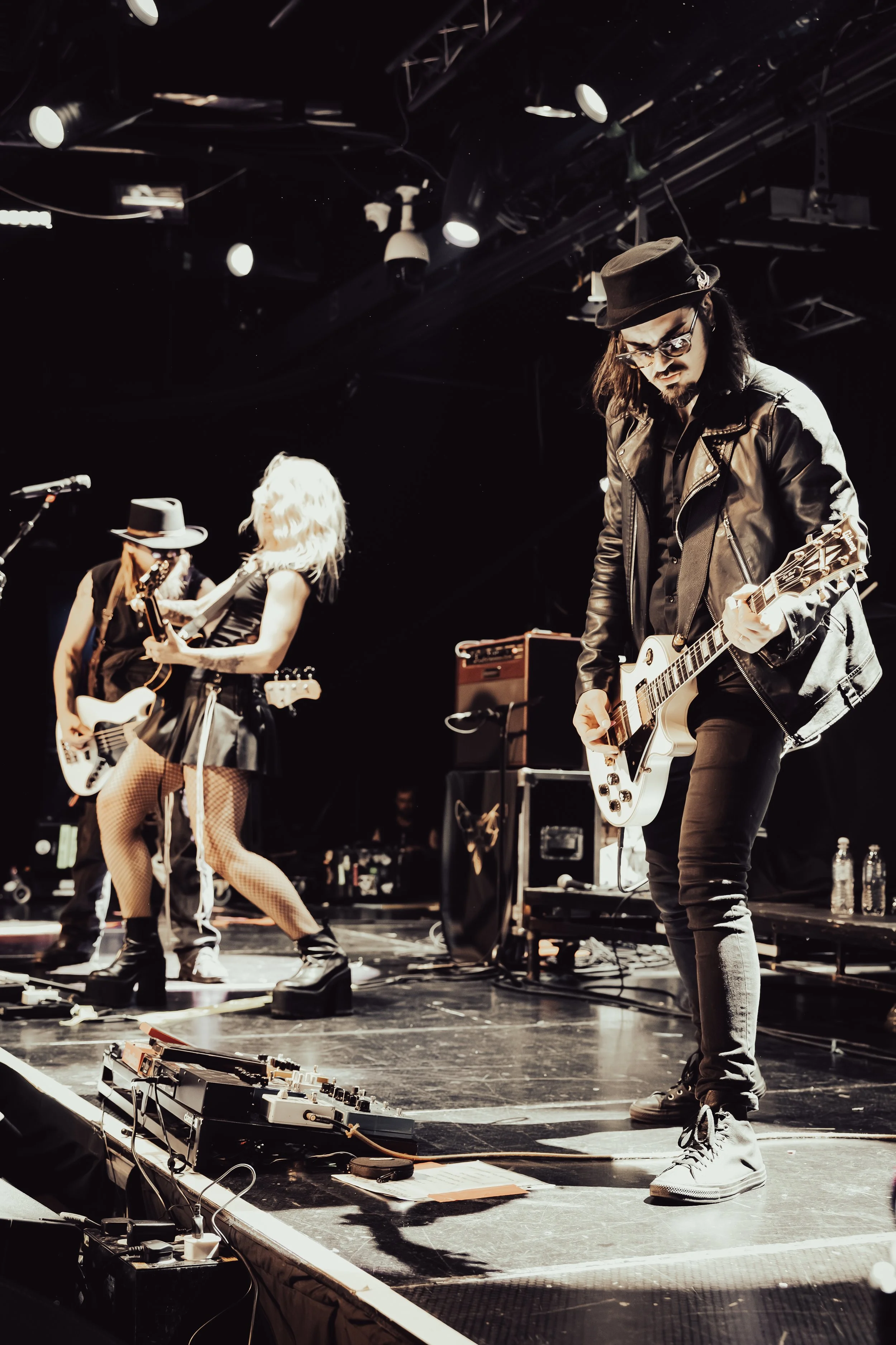 A band performing live on stage, with three musicians visible: a male guitarist wearing glasses, a black hat, and a leather jacket, a female guitarist with long blonde hair, fishnet stockings, and a black dress, and another male guitarist with long h