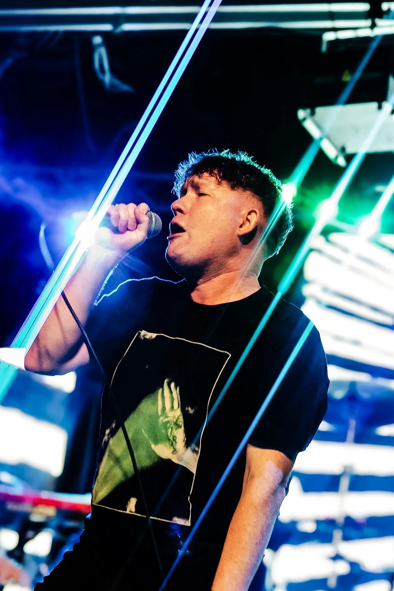 A young man with short curly hair, wearing a black T-shirt with a graphic print, singing into a microphone on stage with bright, colorful neon lights in the background.