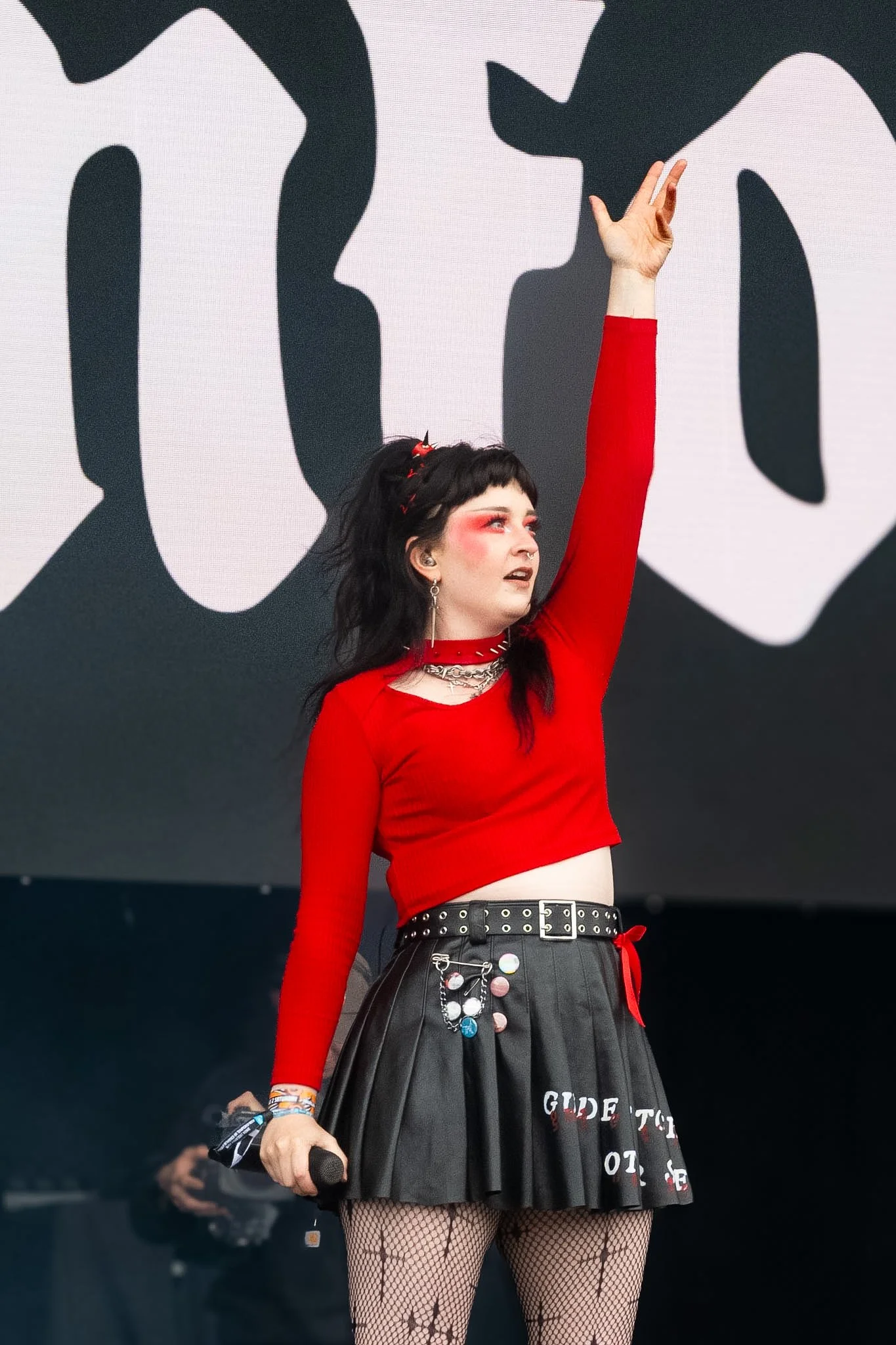 A woman with black hair and red makeup on her eyes and cheeks, wearing a red long-sleeve top, a black pleated skirt with patches and writing, fishnet stockings, and jewelry, holding a microphone, on stage with a black and white abstract background.