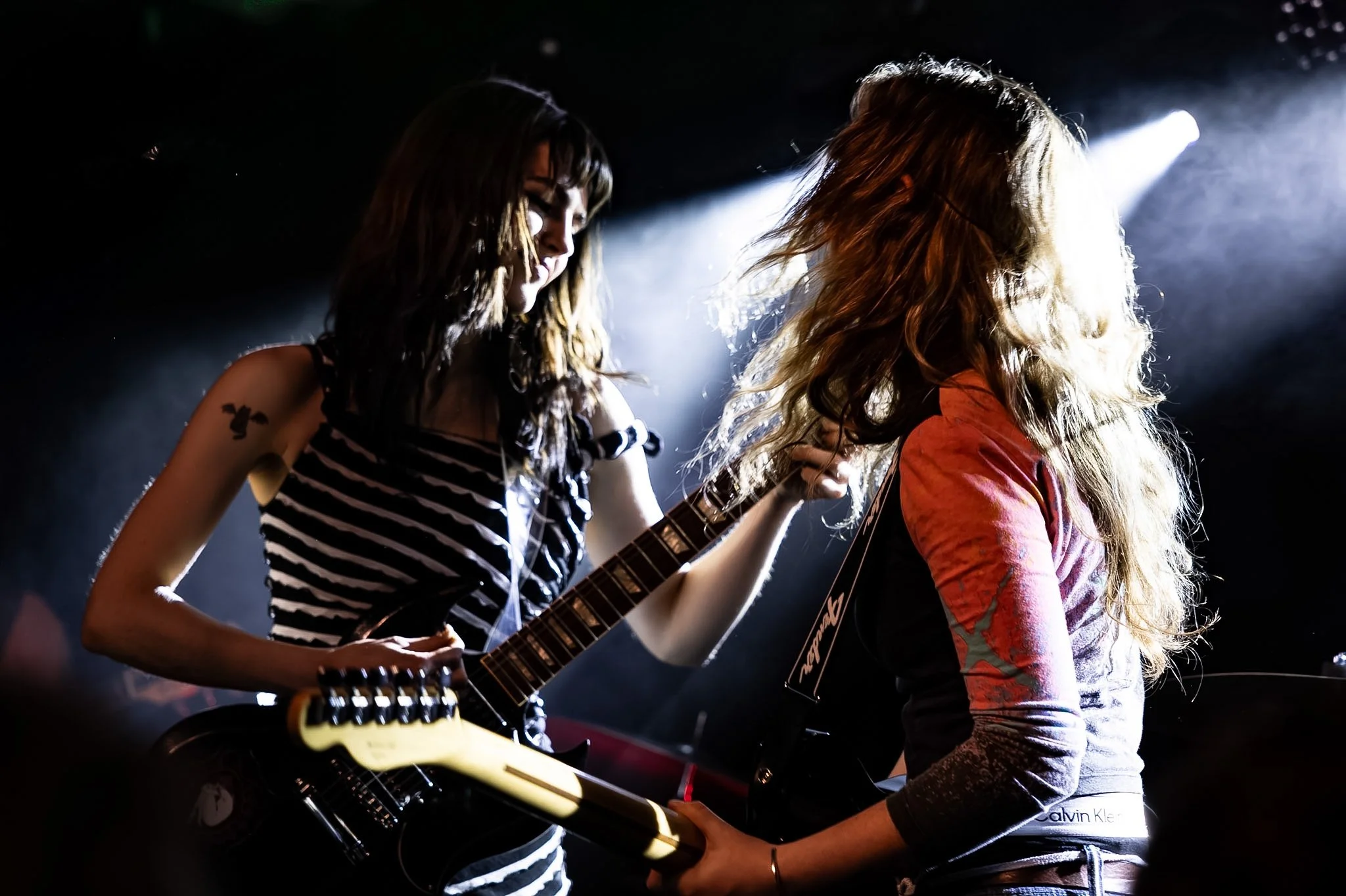 Two women are performing on stage; one is playing an electric guitar and the other is singing or playing an instrument. They have long hair, and stage lights illuminate their backs.