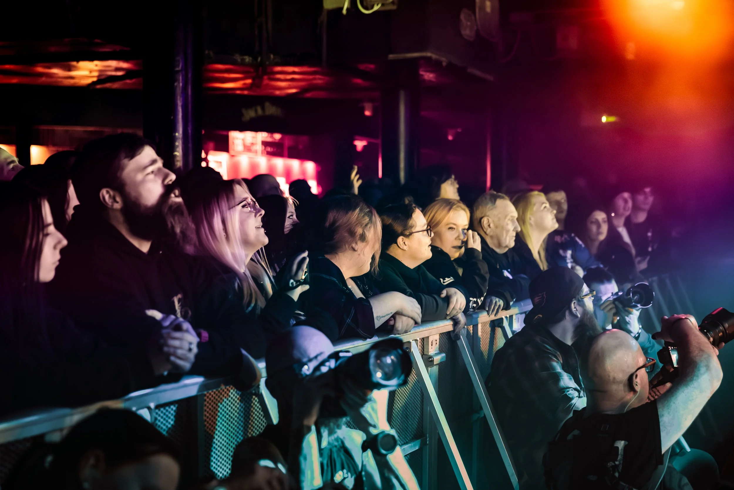 Audience watching a performance or event in a dimly lit indoor venue illuminated with colored lights, with some audience members wearing glasses and capturing the moment with their phones.