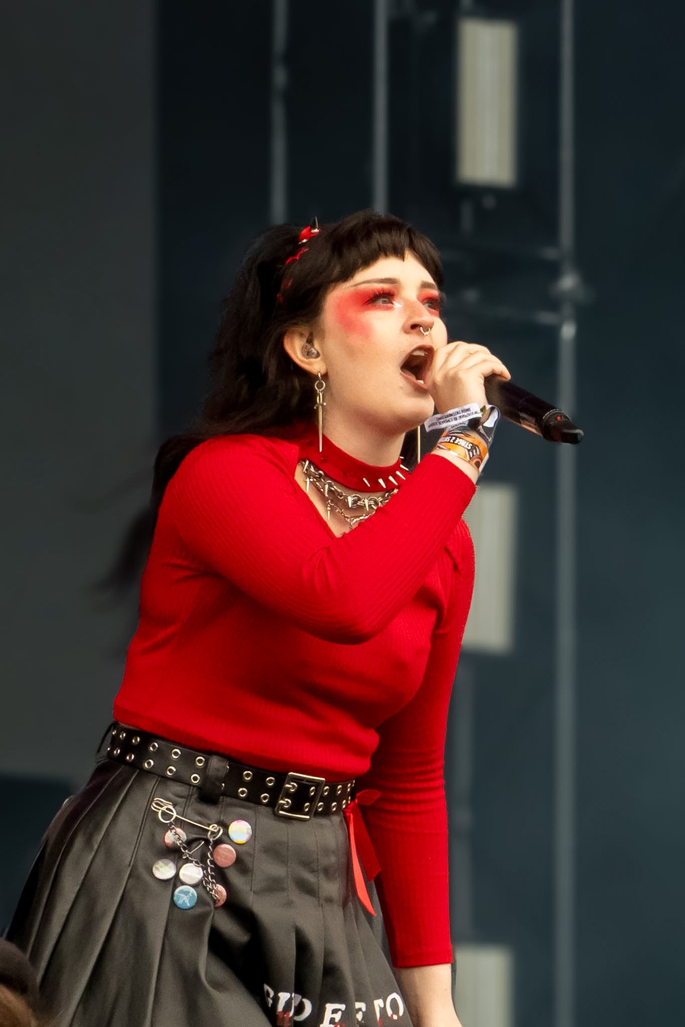 A person with short dark hair, heavy eye makeup, and multiple earrings singing into a microphone. They are wearing a red top, a black pleated skirt decorated with various pins and buttons, and layered necklaces.