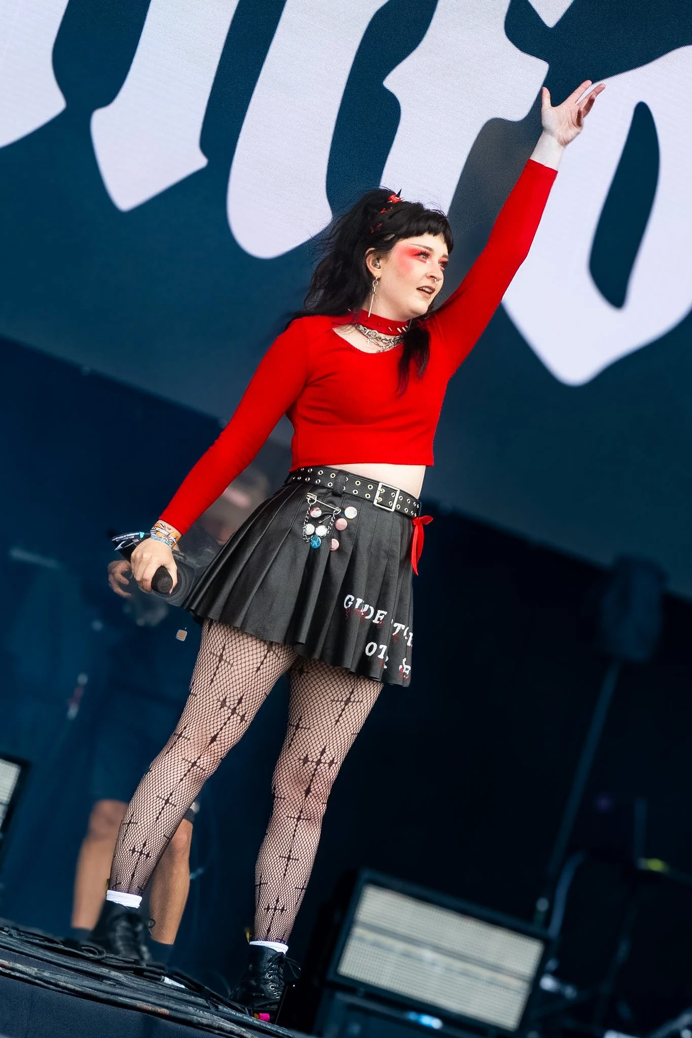 A woman performing on stage, wearing a red long-sleeve crop top, a black pleated skirt with patches and writing, fishnet stockings, and black combat boots. She has dark hair with colorful accessories, dramatic makeup, and is holding a microphone, wit
