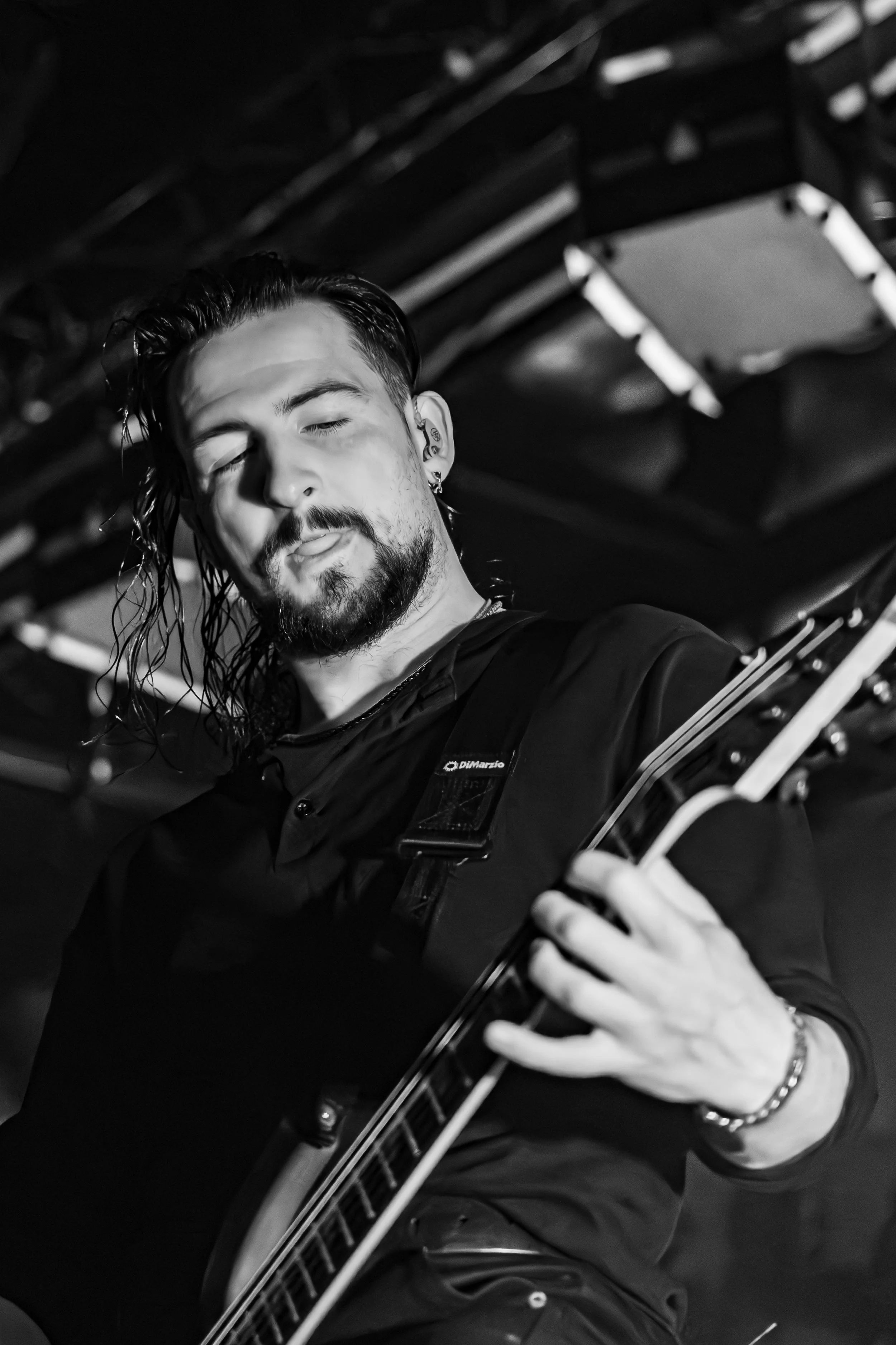 A man with long hair, beard, earrings, and a bracelet playing an acoustic guitar, with his eyes closed and tongue slightly out, in a dark setting.