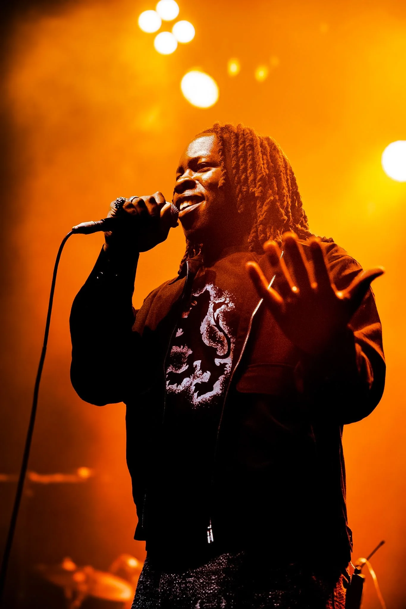 A singer performing on stage under warm orange lighting, holding a microphone, wearing a black jacket and a shirt with a skull print, with dreadlocks and expressive facial expression.