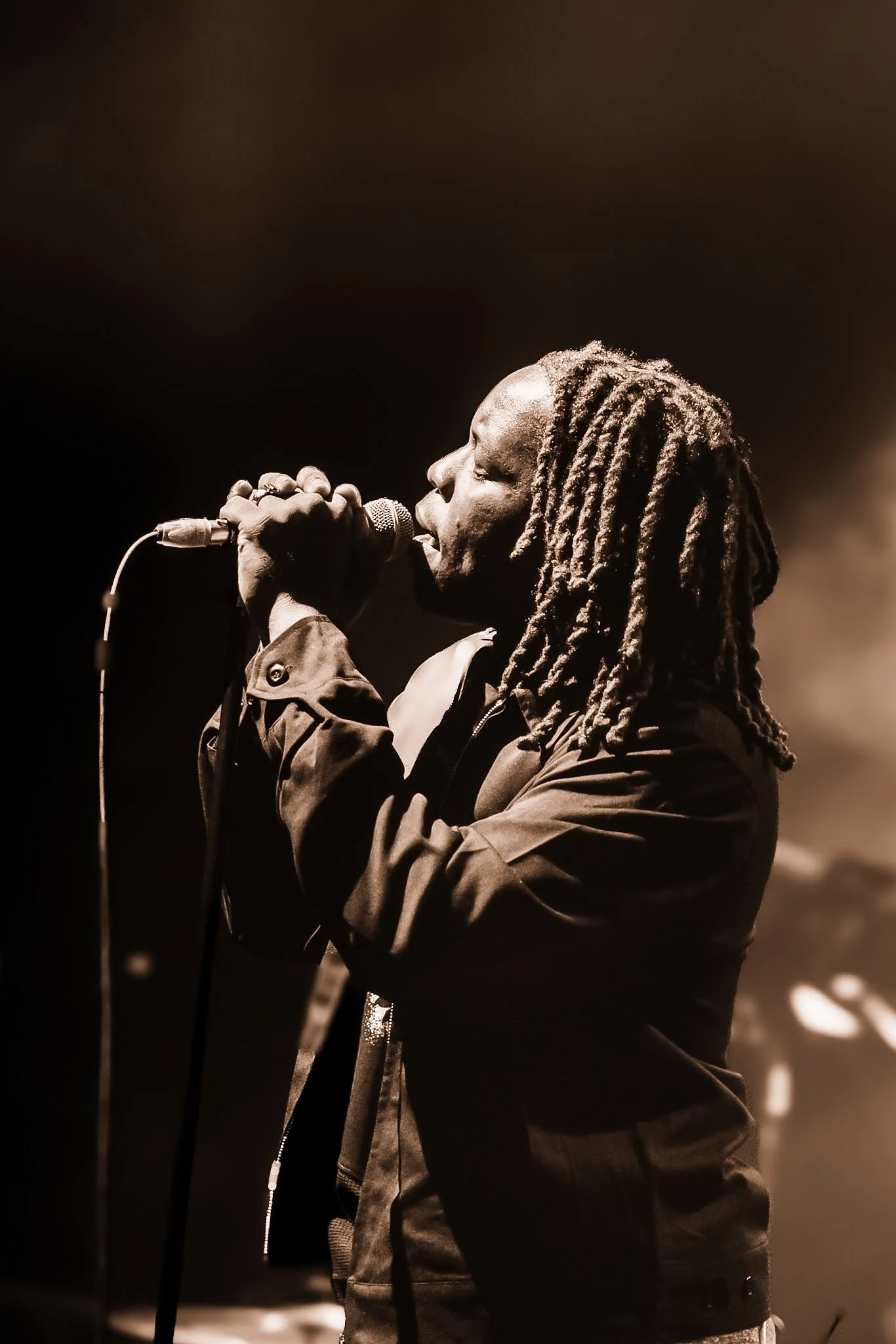 A person with dreadlocks singing into a microphone on stage in a dimly lit setting