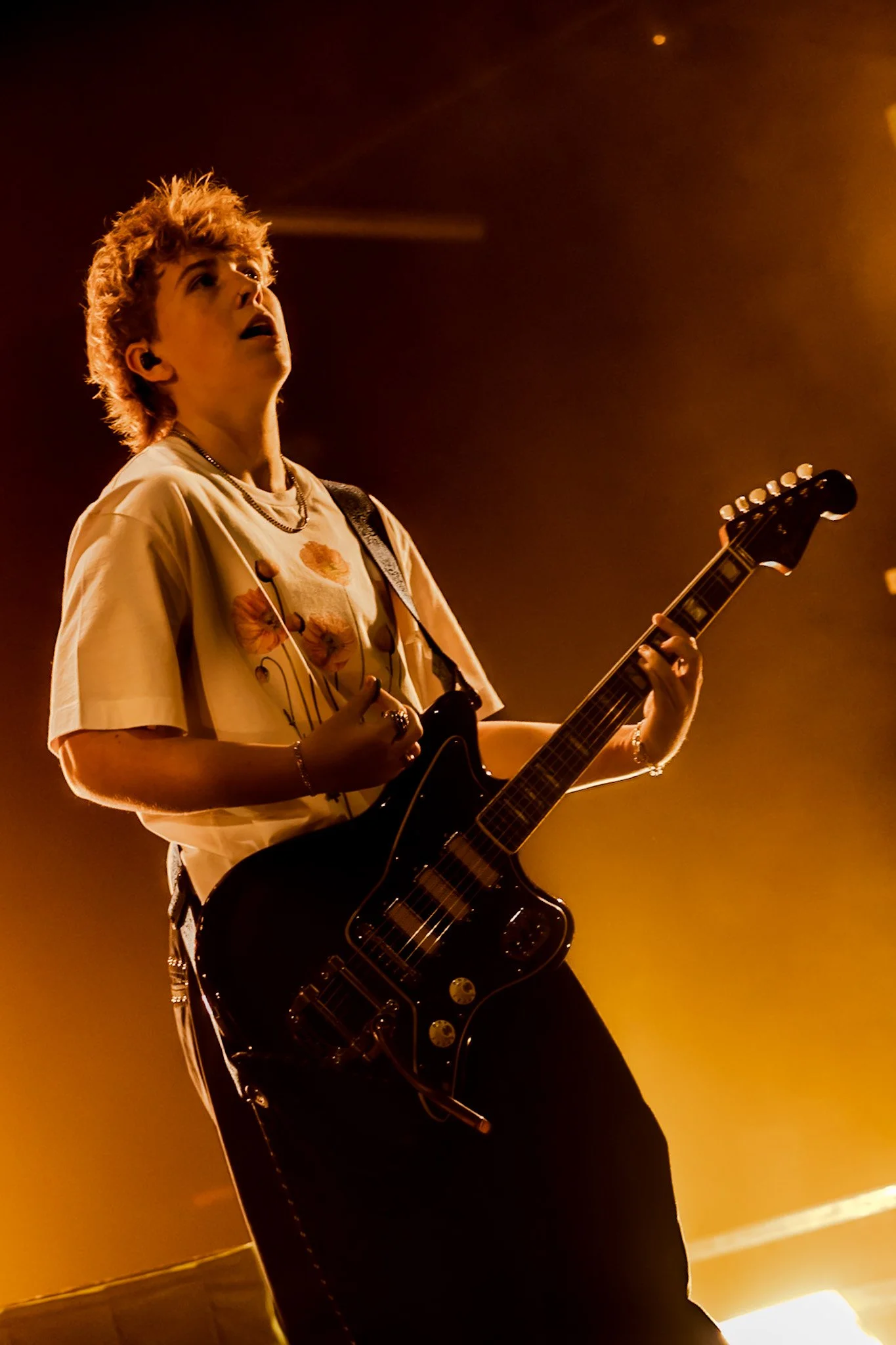 A young person with styled hair wearing a graphic t-shirt, playing an electric guitar on stage with warm, orange lighting creating a dramatic atmosphere.