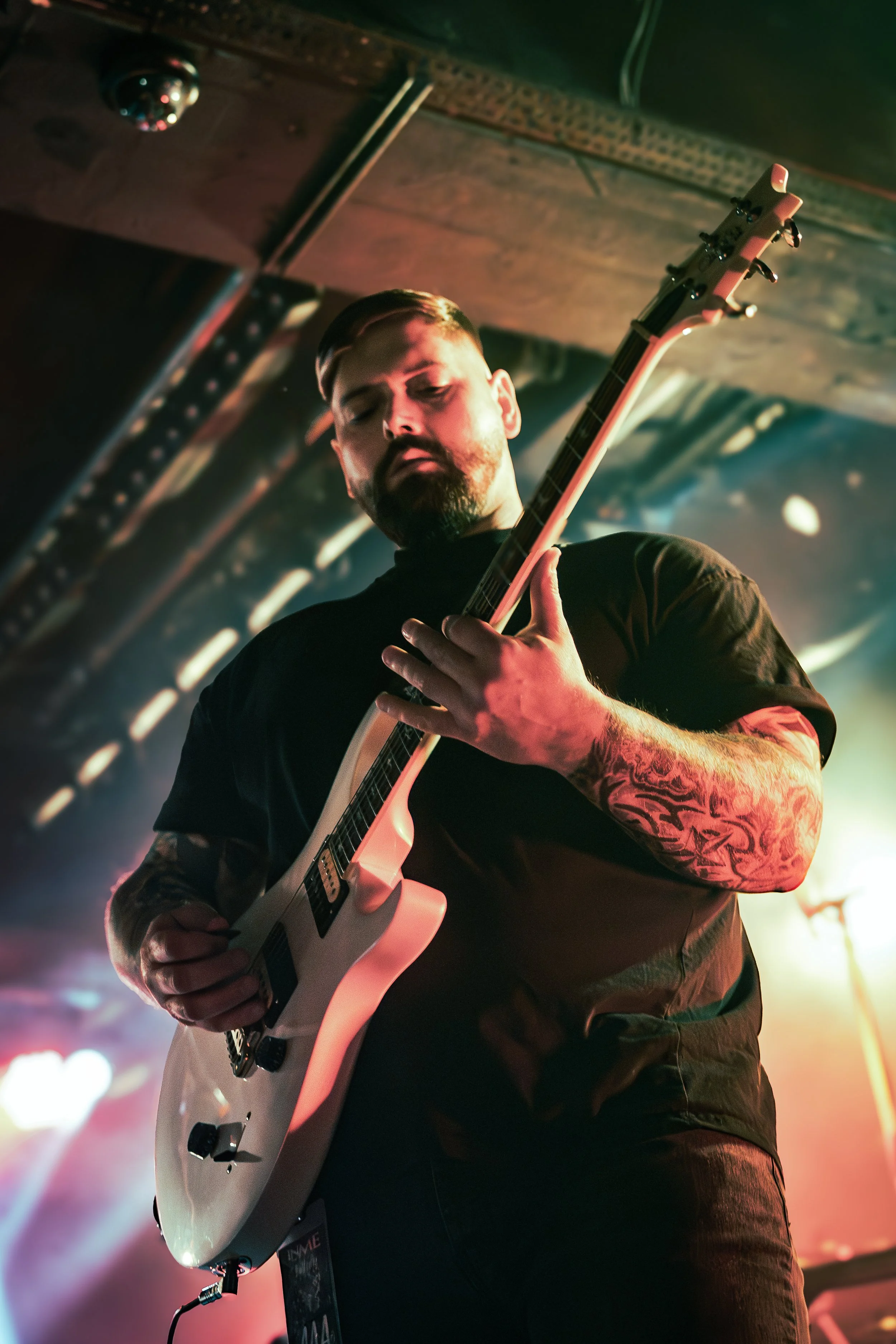 A man with a beard and tattoos on his arm, wearing a black shirt, is playing an electric guitar on stage, with colorful stage lights and a dark background.