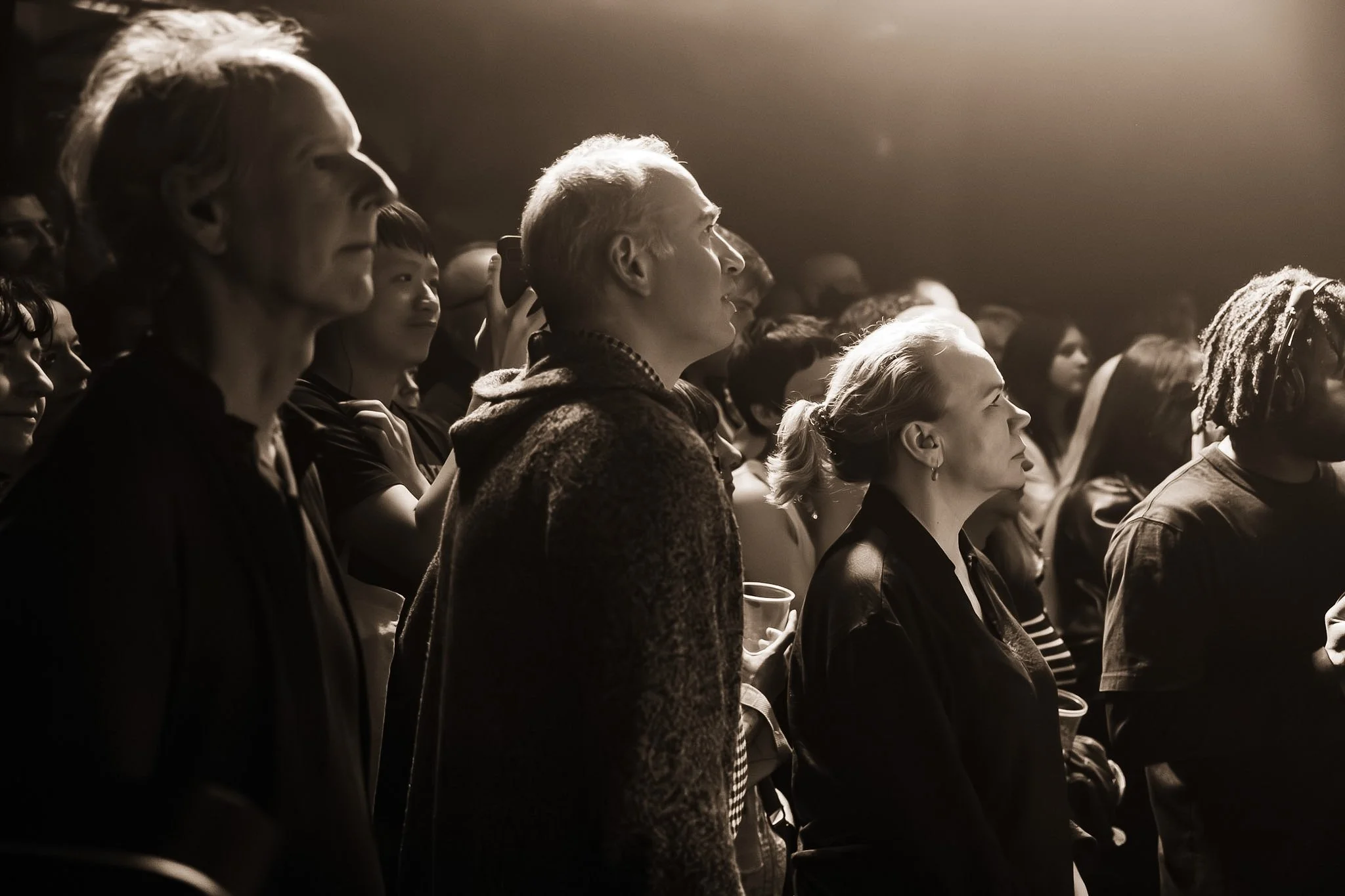 Audience members watching a presentation or performance, all facing forward and illuminated by stage lighting in a dim environment.