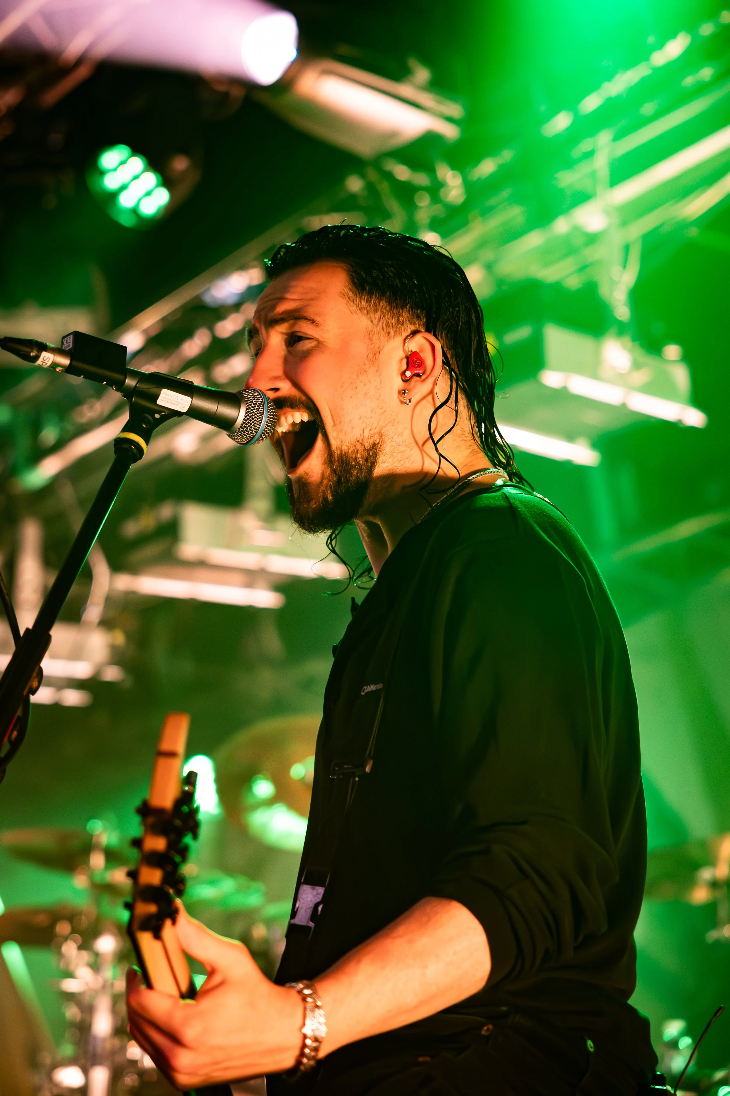 A male musician singing into a microphone at a live concert, with green stage lighting and blurred drum set in the background.