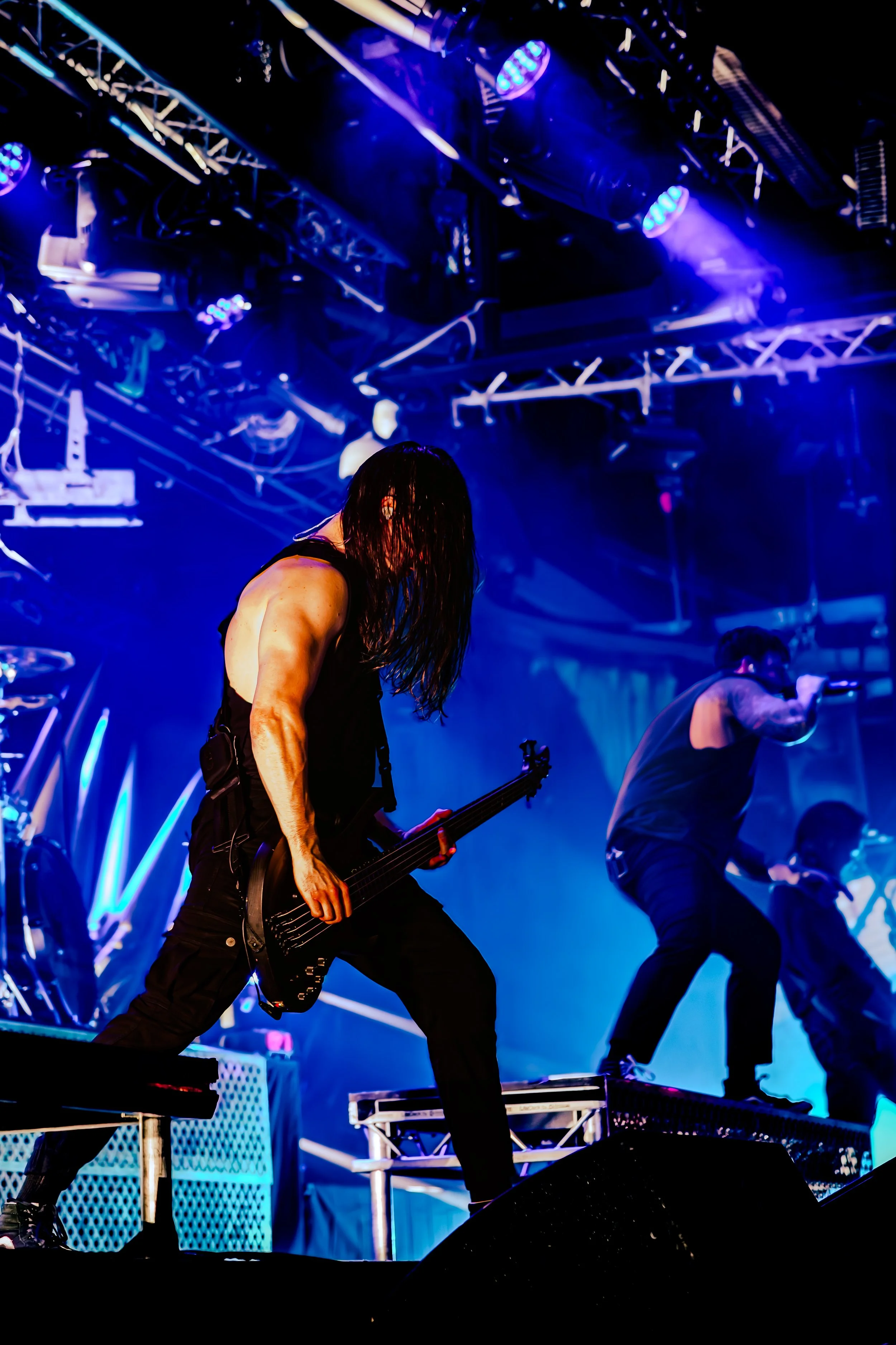 A live rock band performing on stage with blue dramatic lighting, featuring a guitarist in the foreground with long dark hair, wearing a sleeveless top, and a bassist and vocalist in the background.