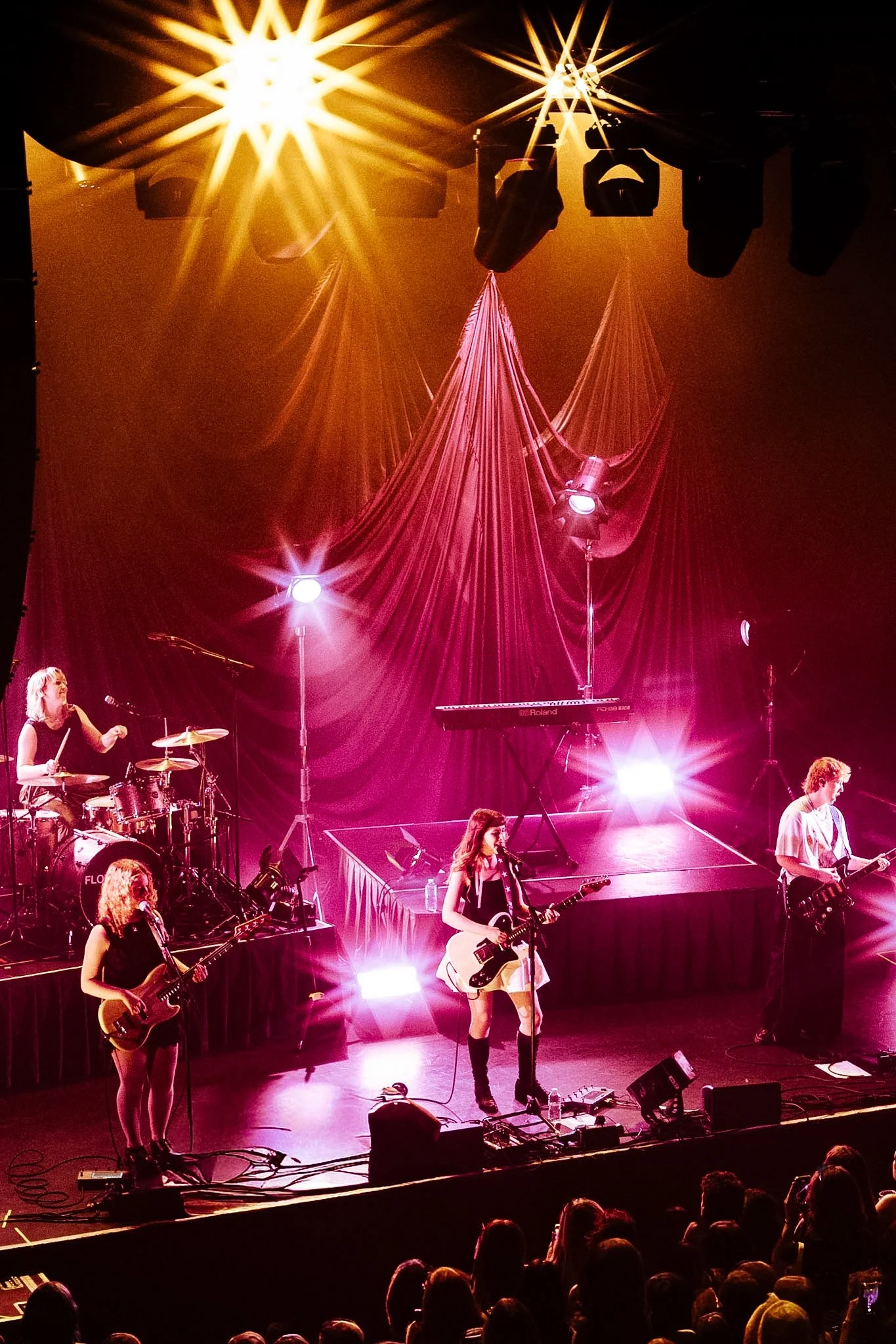 A live music performance with multiple musicians on stage. There are five women with guitars, a drummer, and a keyboard player. The stage is decorated with draped fabric and lighting effects, including pink and yellow lights, creating a vibrant atmos