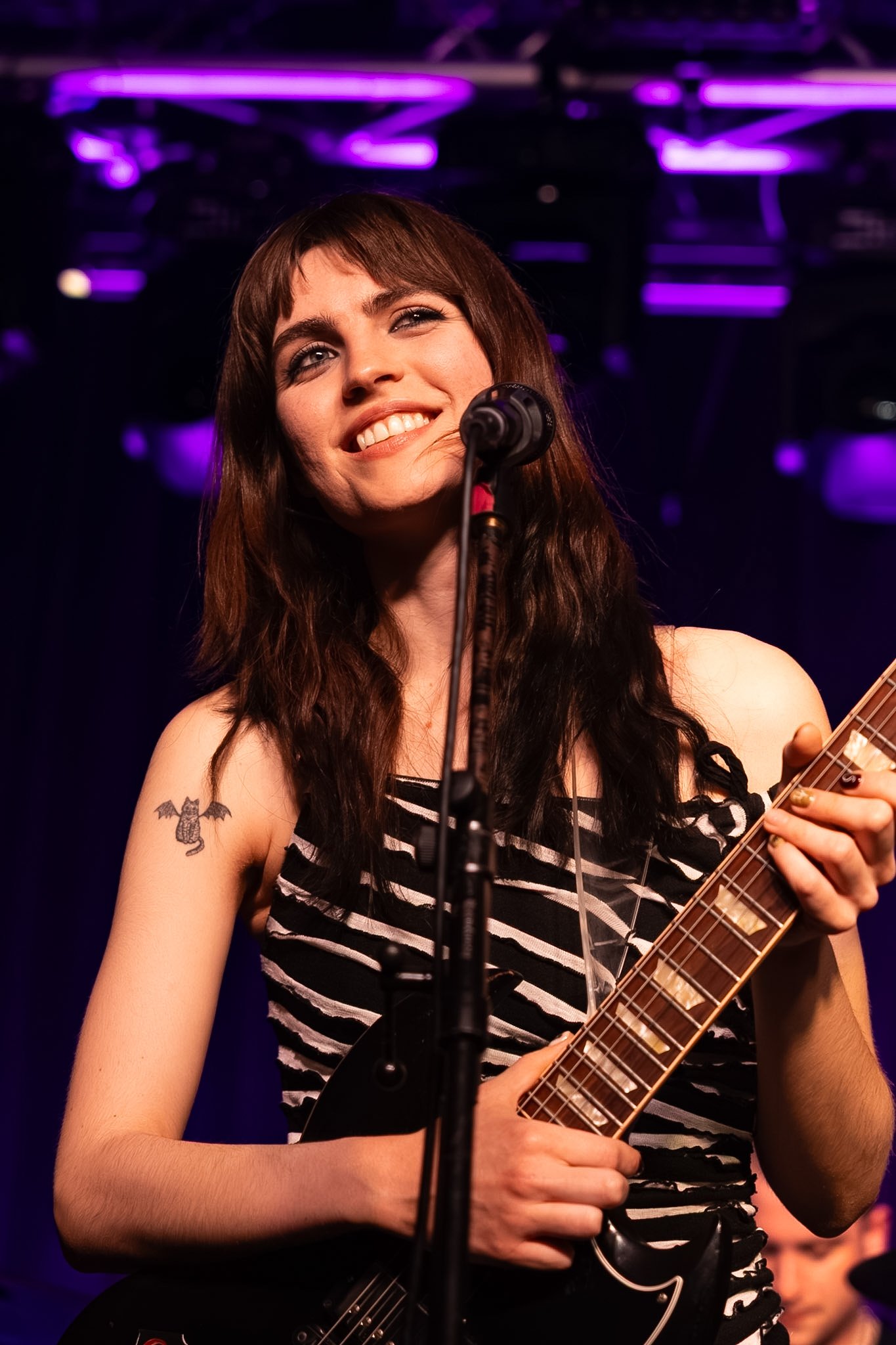 A young woman with brown hair and a tattoo of a bat on her arm, smiling while holding a guitar on stage with purple lighting overhead.