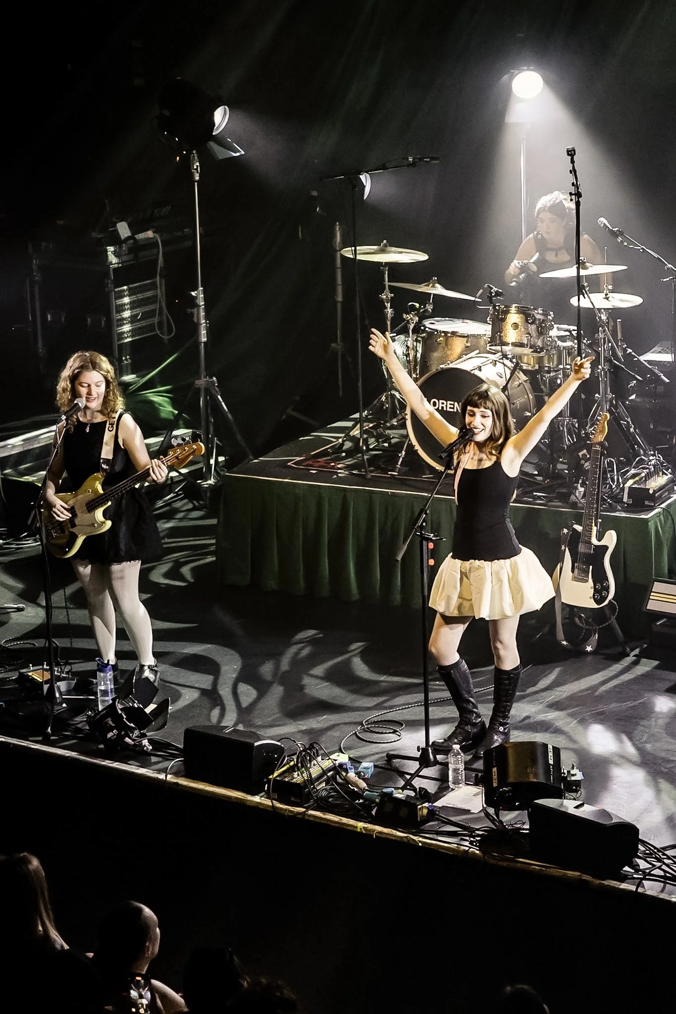 Two female singers perform on stage with musical instruments, including a bass guitar and a drum set, under stage lights during a concert.