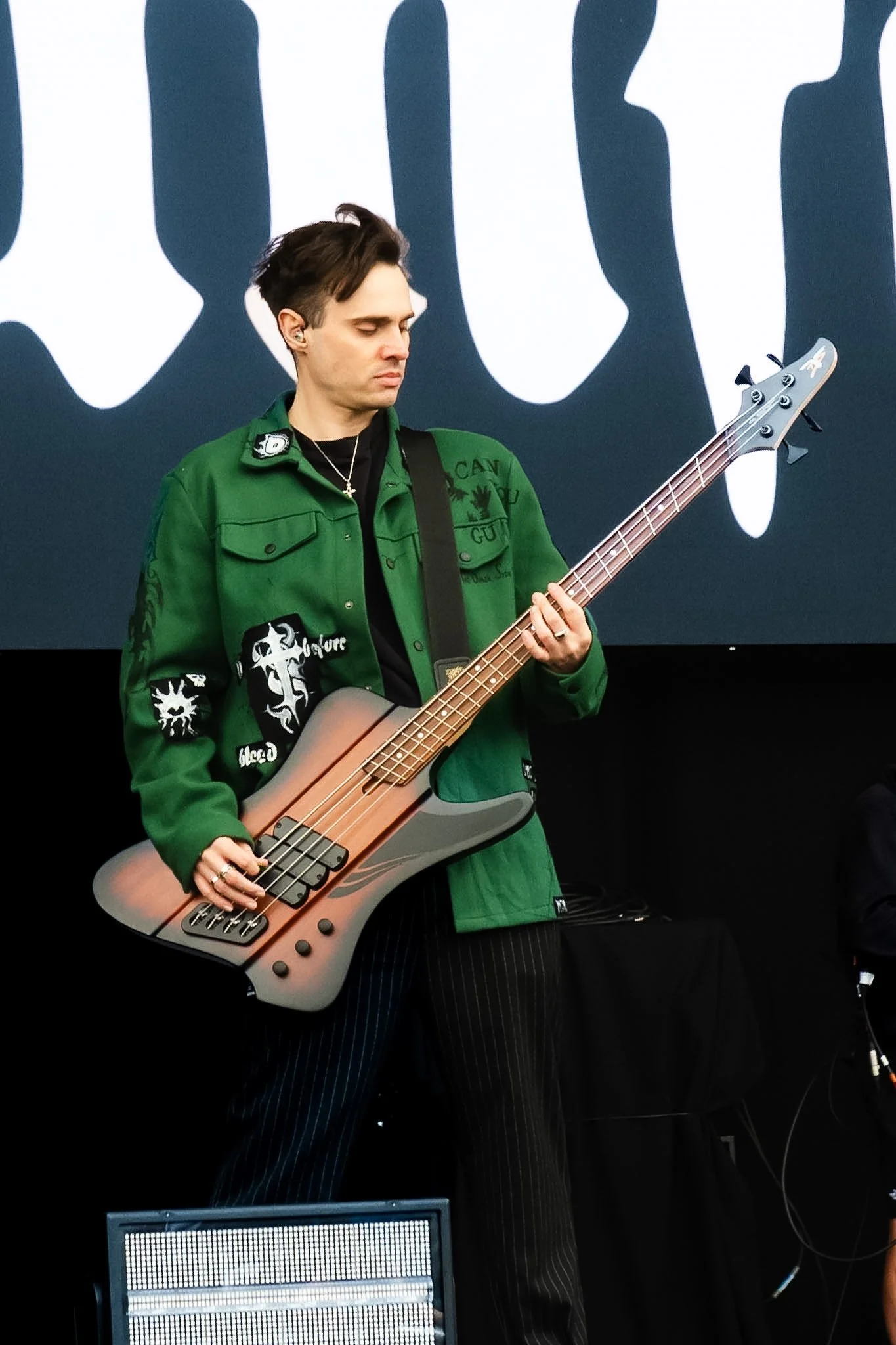 A young man with dark hair playing a bass guitar on stage, wearing a green jacket with black and white patches, and striped black pants.