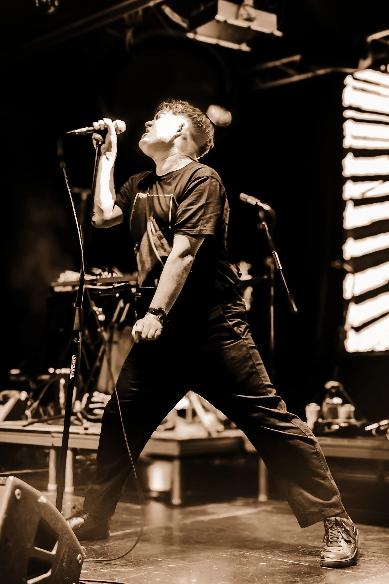 A male singer on stage passionately singing into a microphone with a dynamic pose, legs spread apart, head tilted back, in a dark performance setting with stage lighting and equipment visible in the background.