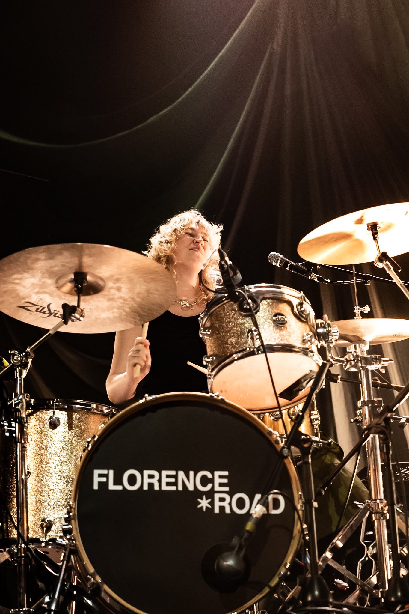 A female drummer with blonde curly hair playing a drum set on stage, illuminated by a warm overhead light. The drum kit includes a bass drum with the words 'FLORENCE ROAD' printed on it, and cymbals labeled 'Zildjian'.