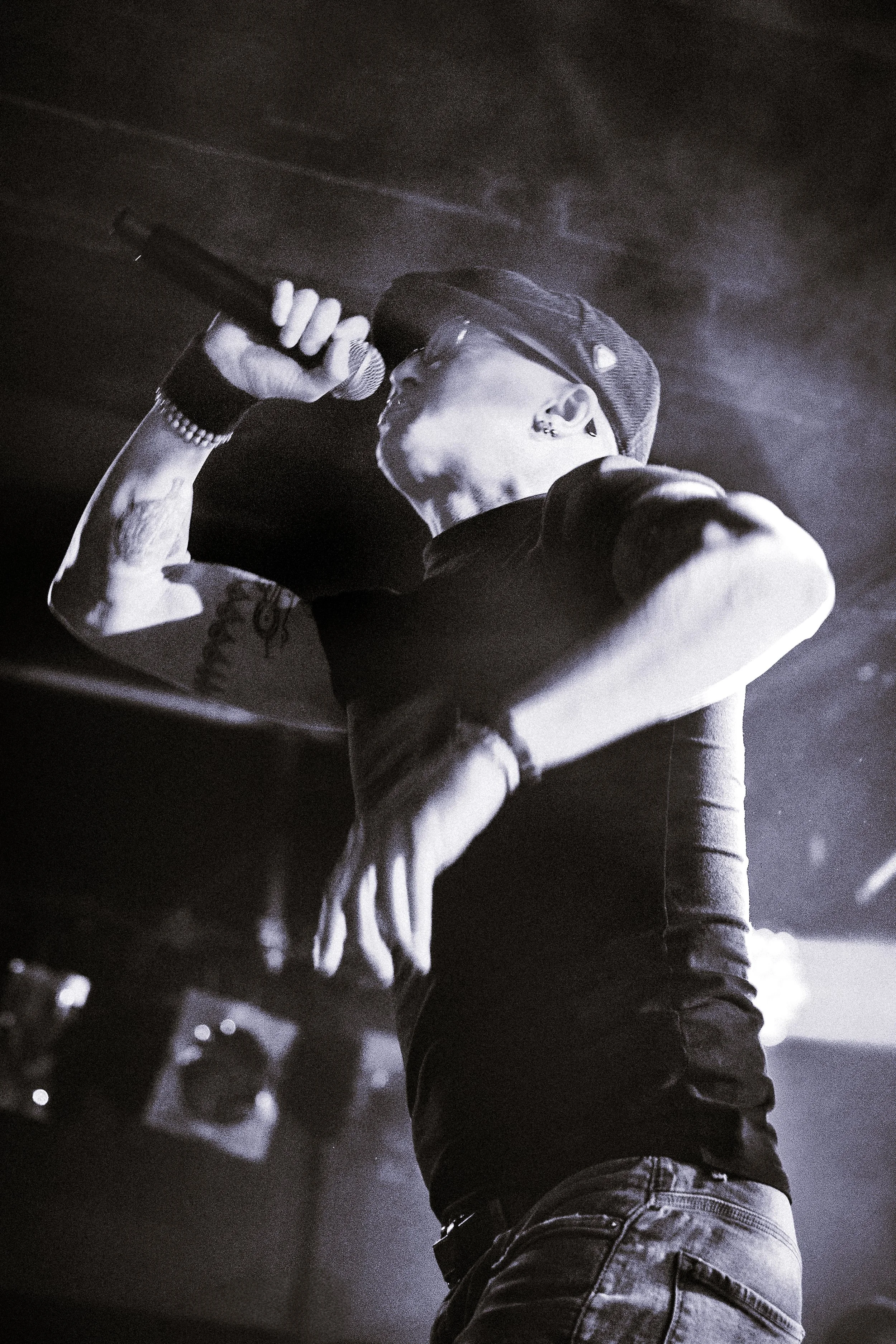 A person singing into a microphone on stage, wearing glasses, earrings, a cap, and a wristband, with tattoos visible on their arm, in a low-light setting.