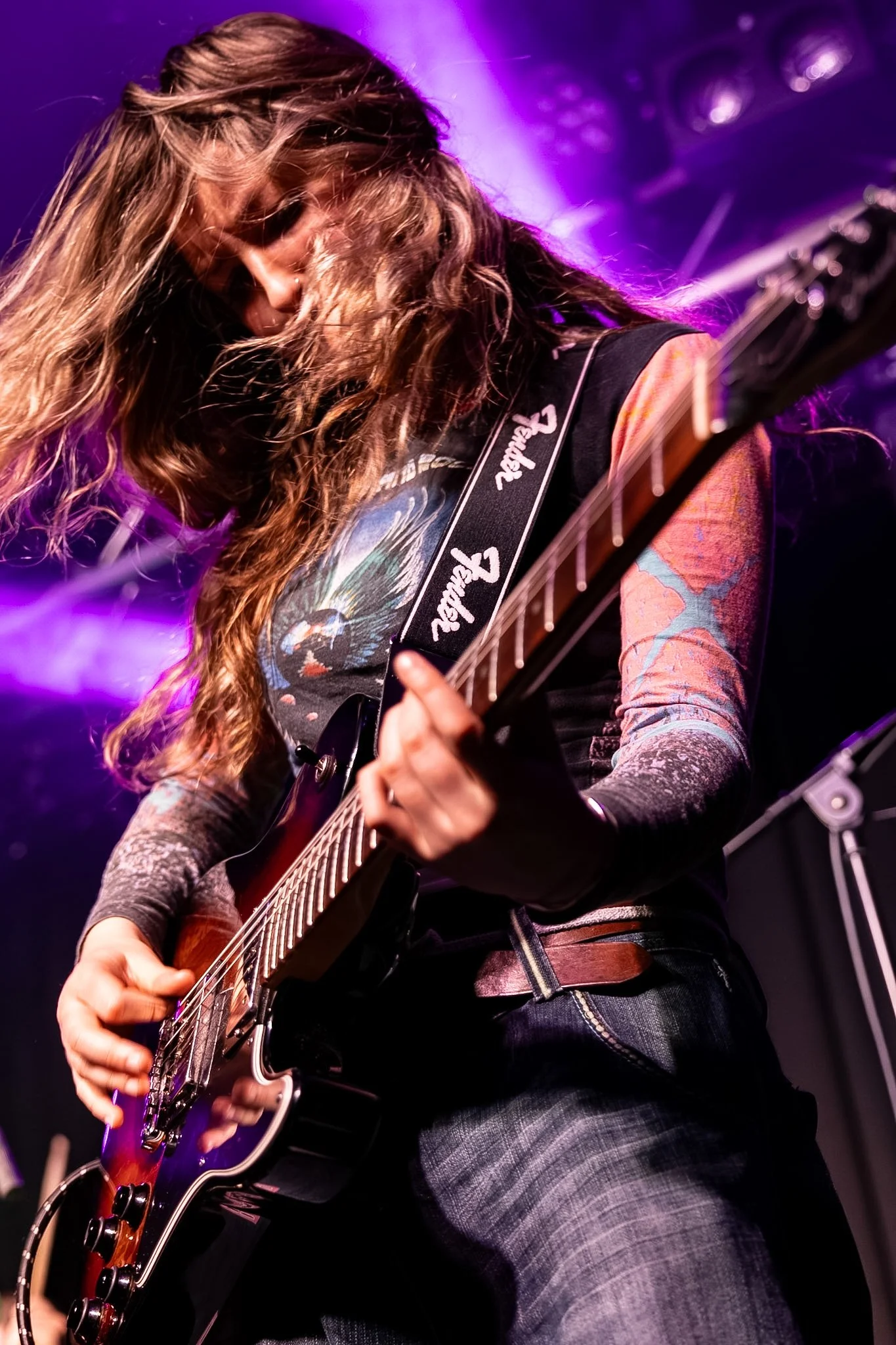 A musician with long wavy hair playing an electric guitar on stage illuminated with purple and blue lights.
