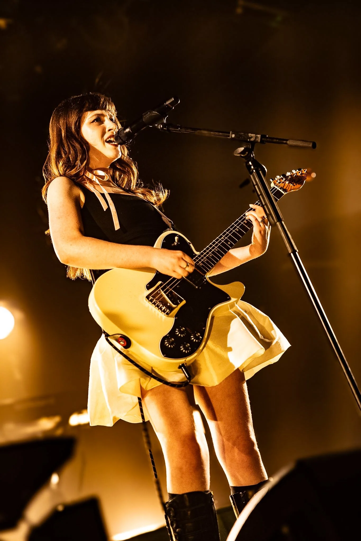 A woman with wavy brown hair singing into a microphone while playing an electric guitar on stage, under warm stage lighting.