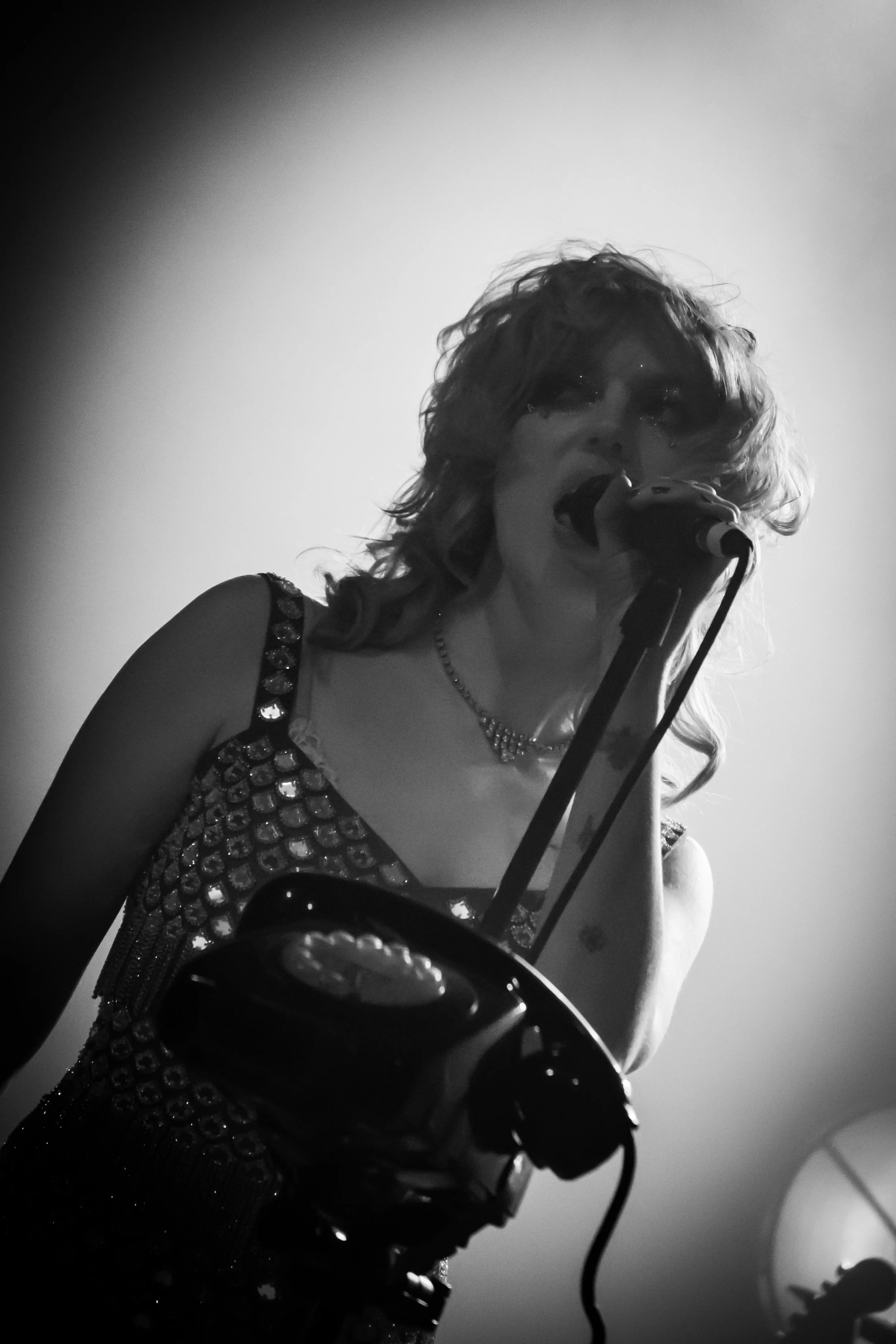 Black and white photo of a woman singing into a microphone, wearing a sparkly dress and jewelry, with a blurry background.