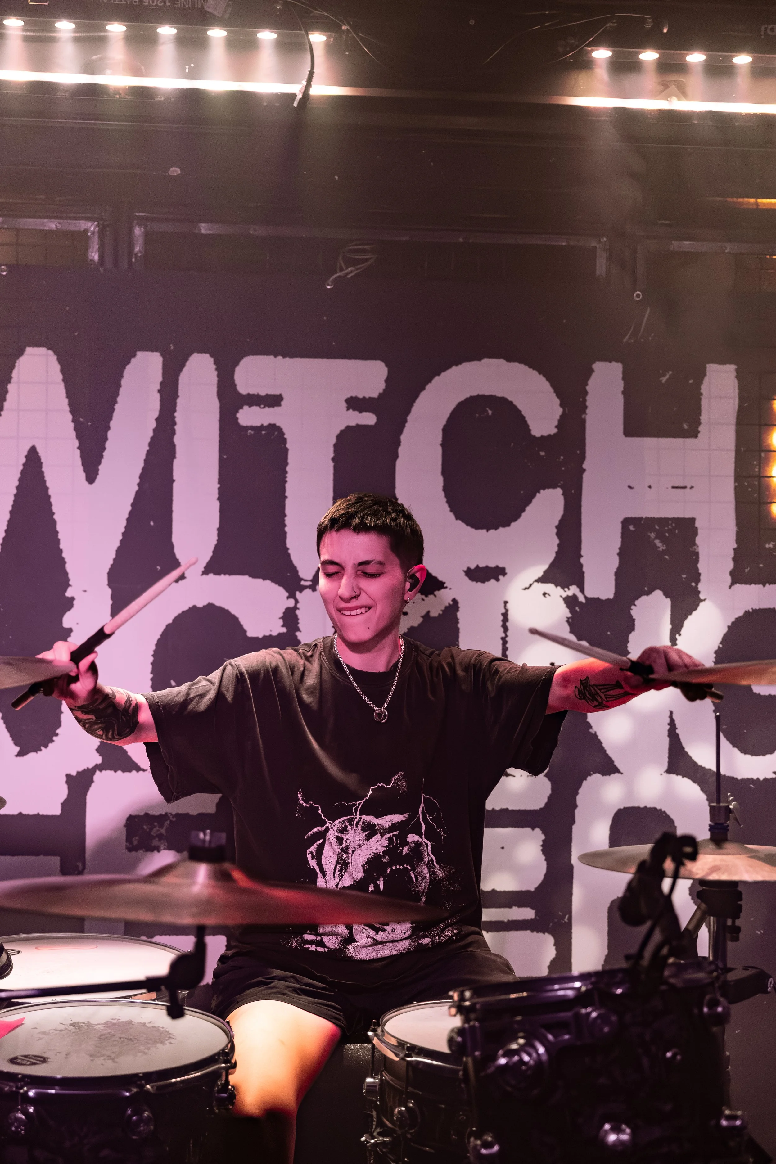 A young person with short dark hair playing drums on stage, wearing a black t-shirt with lightning graphics, with a background that has large white text on a black wall and stage lighting overhead.