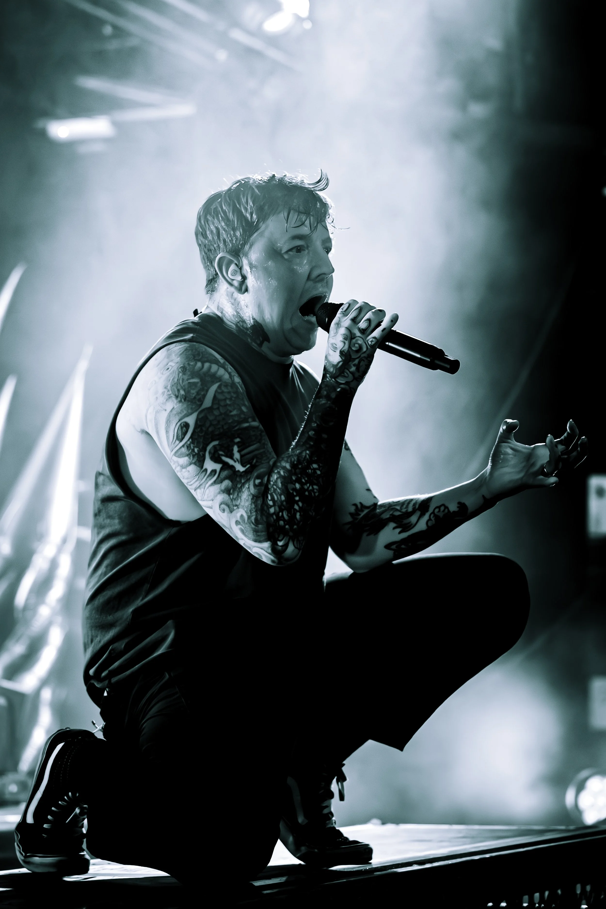 A singer with tattoos, wearing a sleeveless shirt and sneakers, crouching on one knee while holding a microphone during a live performance, with dramatic stage lighting and smoke in the background.