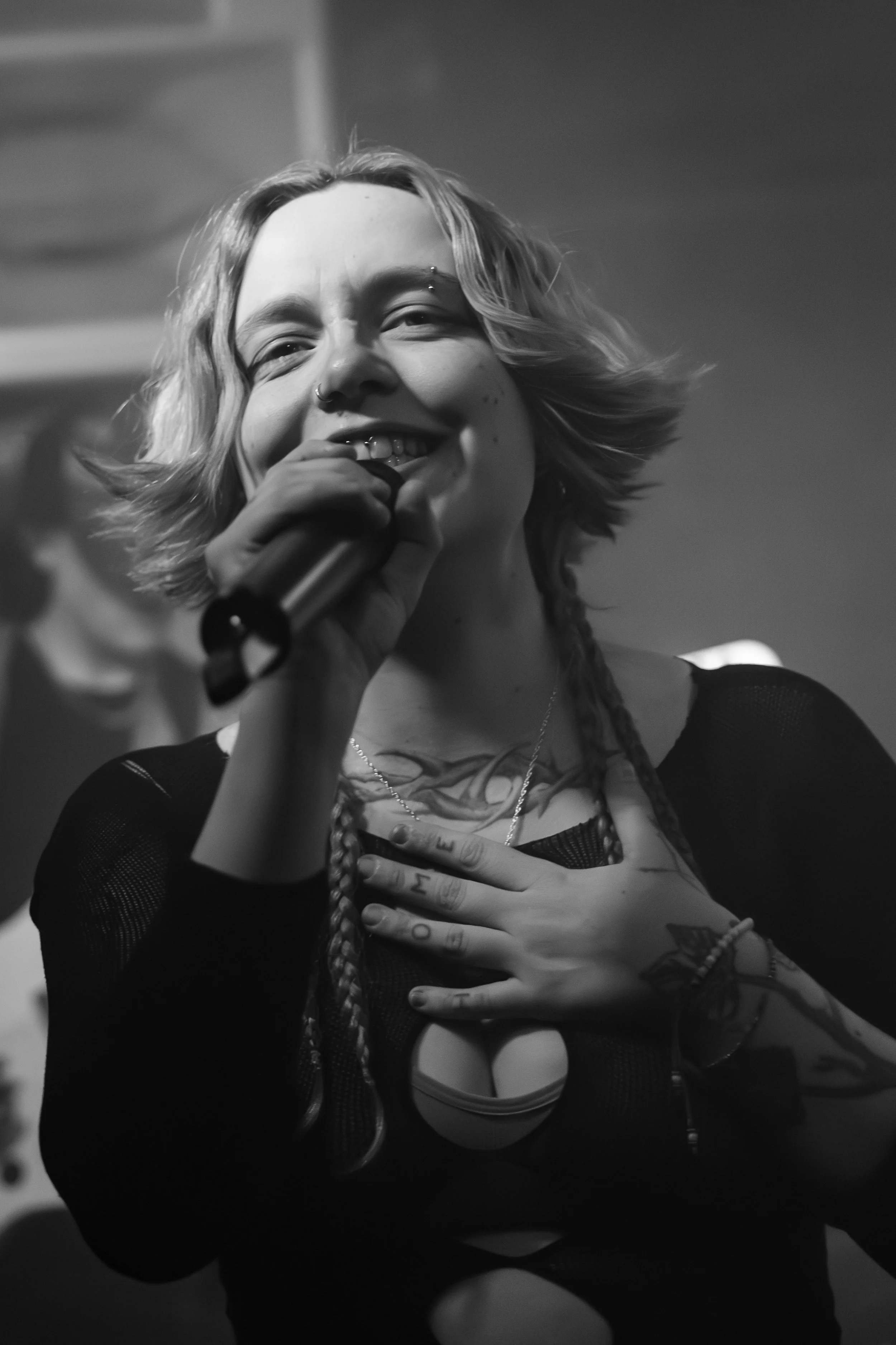A young woman with short, wavy hair and multiple facial piercings is holding a microphone close to her mouth, smiling, and has her left hand on her chest with tattoos on her fingers and wrist. She is dressed in a black mesh top and a tank top underne