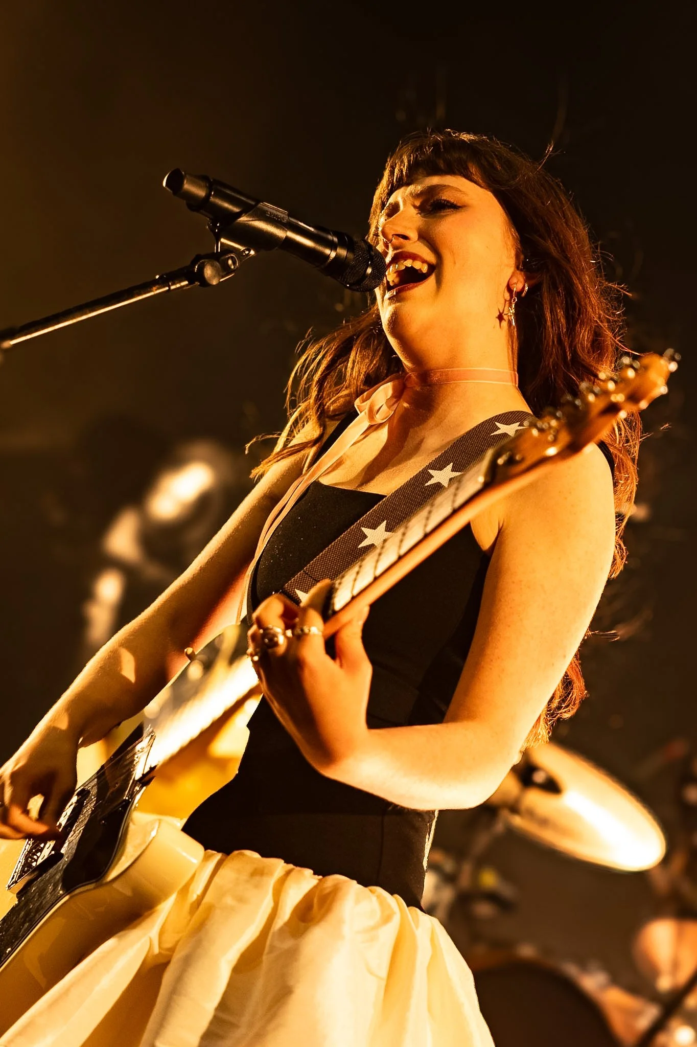 Musician singing into a microphone while playing guitar on stage, dressed in a black top and a yellow skirt, with a pink choker and star-patterned guitar strap, in a warm, dimly lit performance setting.